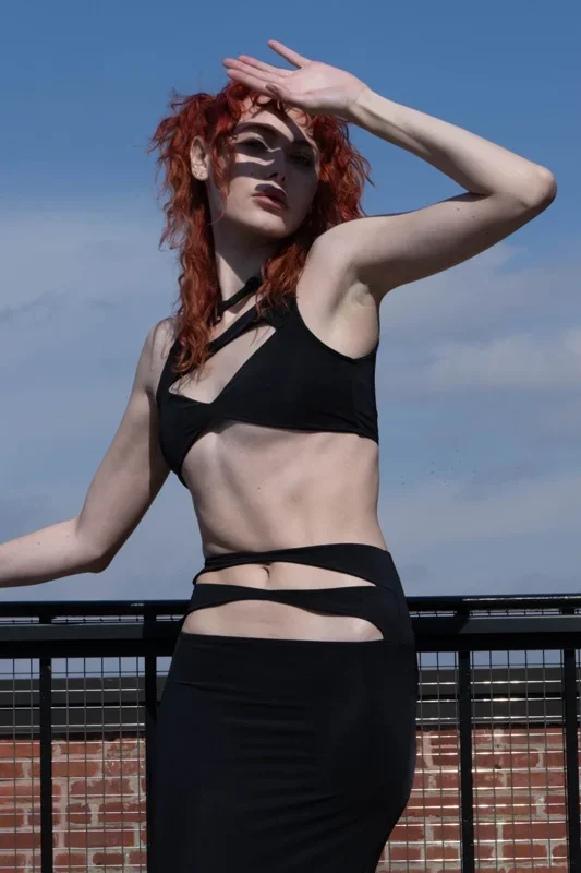 Woman with red curly hair wearing a black cut-out top and skirt standing outdoors on a rooftop with a brick wall, against a partly cloudy sky.