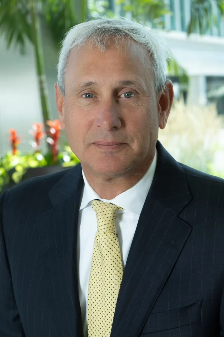 A middle-aged man with gray hair and blue eyes wearing a dark pinstripe suit, white shirt, and yellow tie, standing outdoors with blurred greenery in the background.