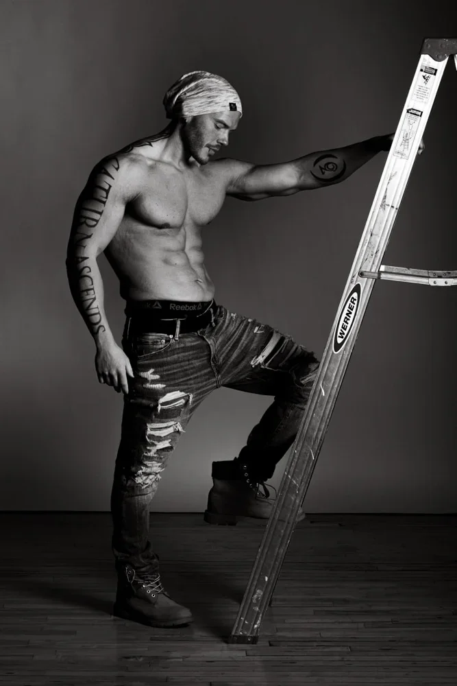 A shirtless man with tattoos and a head wrap stands on a wooden floor, holding onto a stepladder with one foot on the bottom rung and looking down.