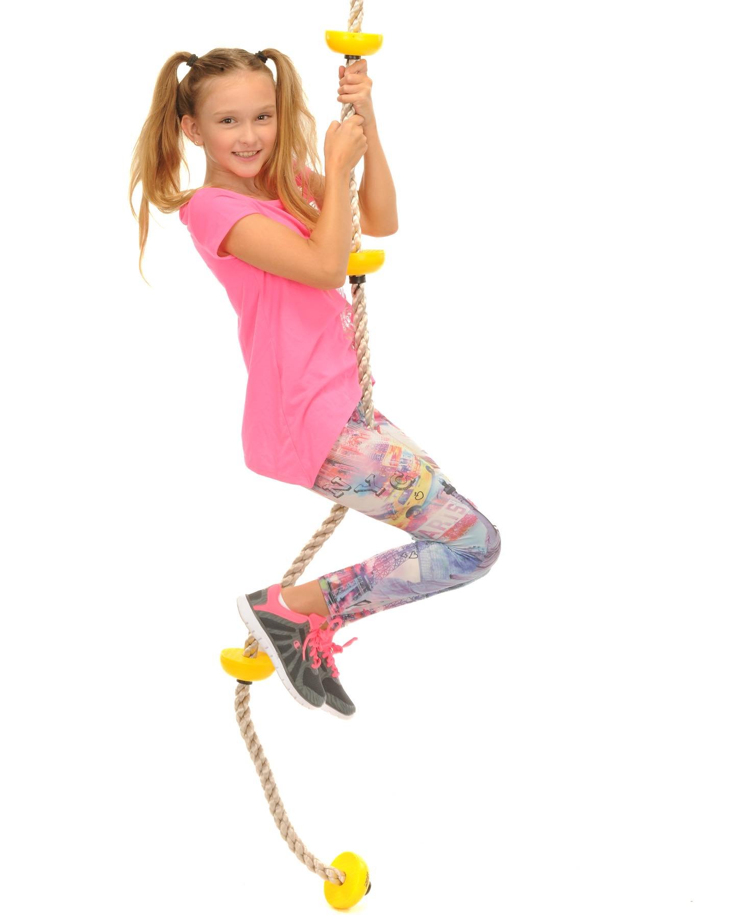 A young girl with blonde hair in pigtails, wearing a pink shirt, colorful leggings, and running shoes, is smiling while sitting on a rope swing with yellow supports, isolated against a white background.