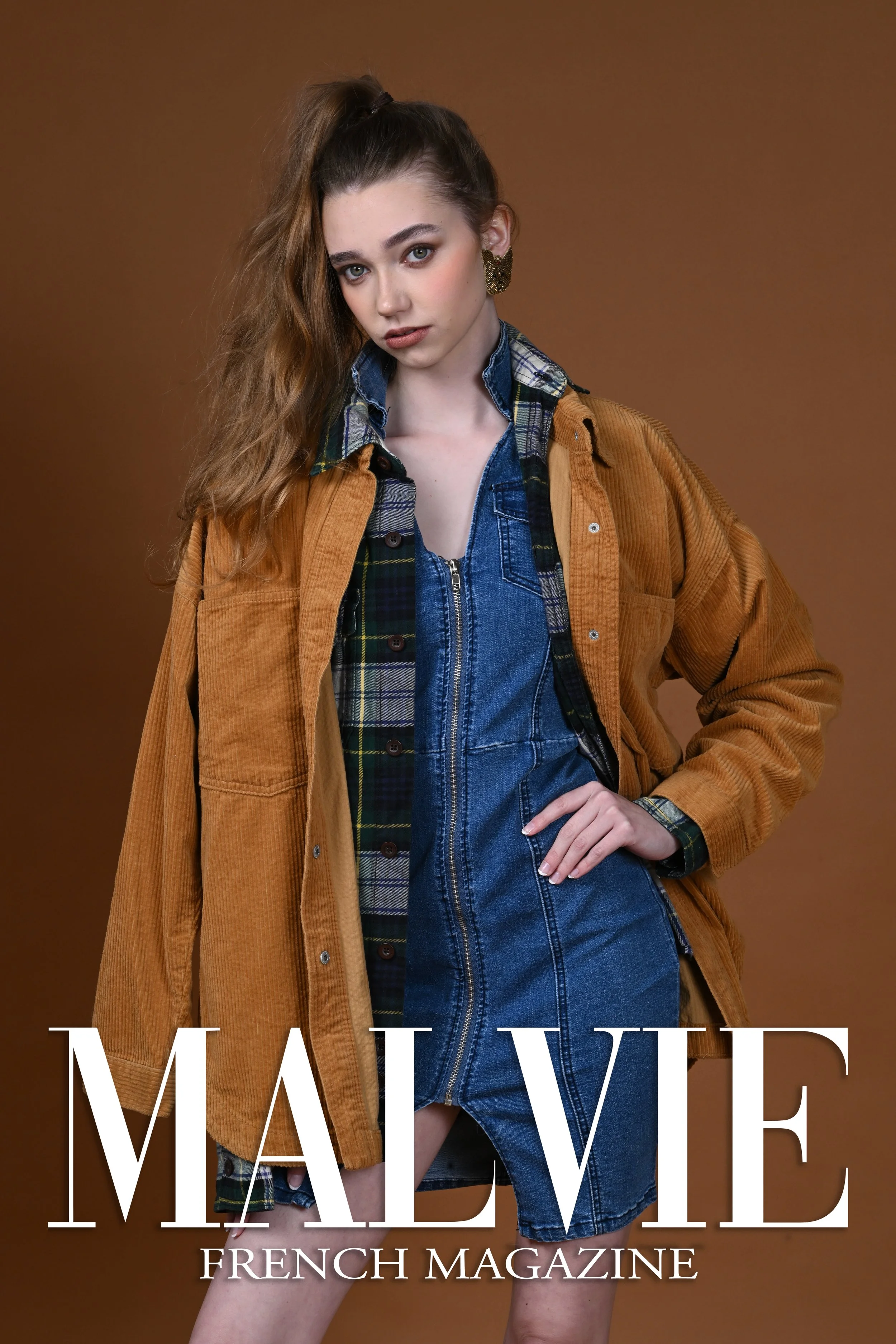 A young woman with long wavy brown hair wearing a layered outfit consisting of a plaid shirt, denim dress, and a brown jacket, standing against a solid brown background, looking directly at the camera, with magazine text overlay reading "MALVIE FRENC