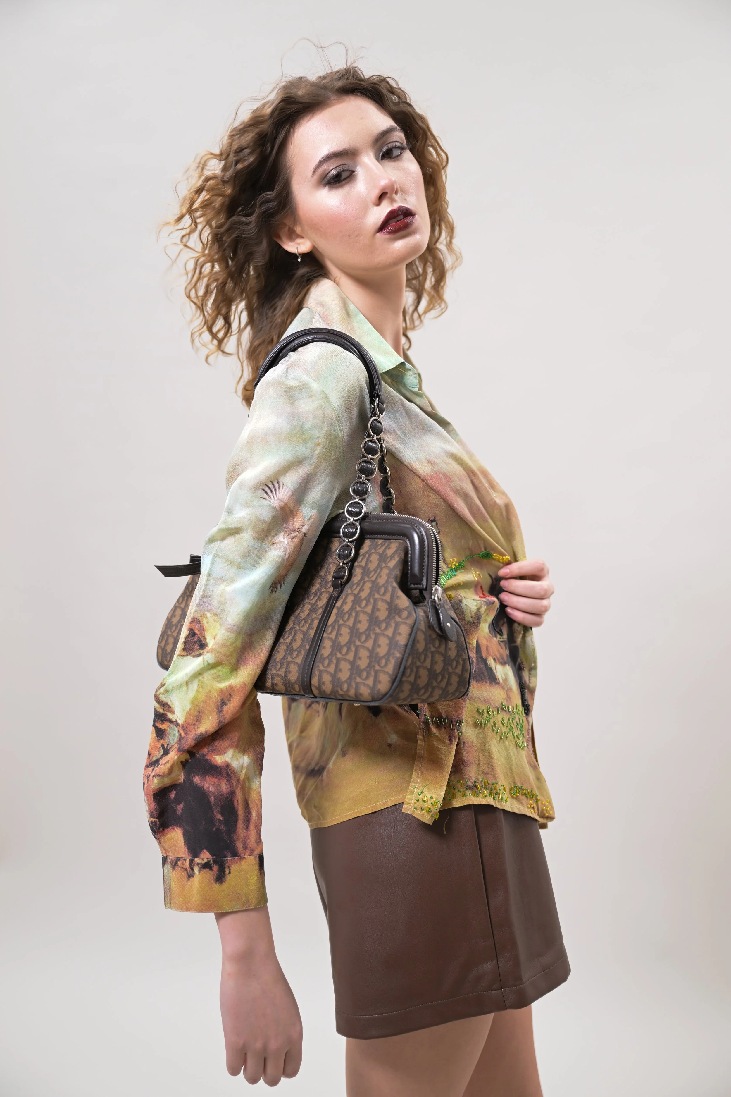 A young woman with curly hair and dark lipstick wearing a floral satin blouse, a brown leather skirt, and carrying a designer handbag with a chain strap against a plain background.