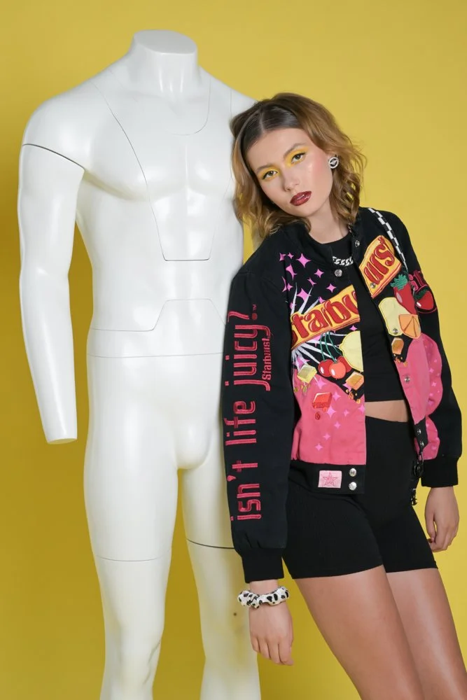 Young woman with brown wavy hair and makeup wearing a colorful graphic jacket and black shorts, standing next to a white mannequin against a yellow background.