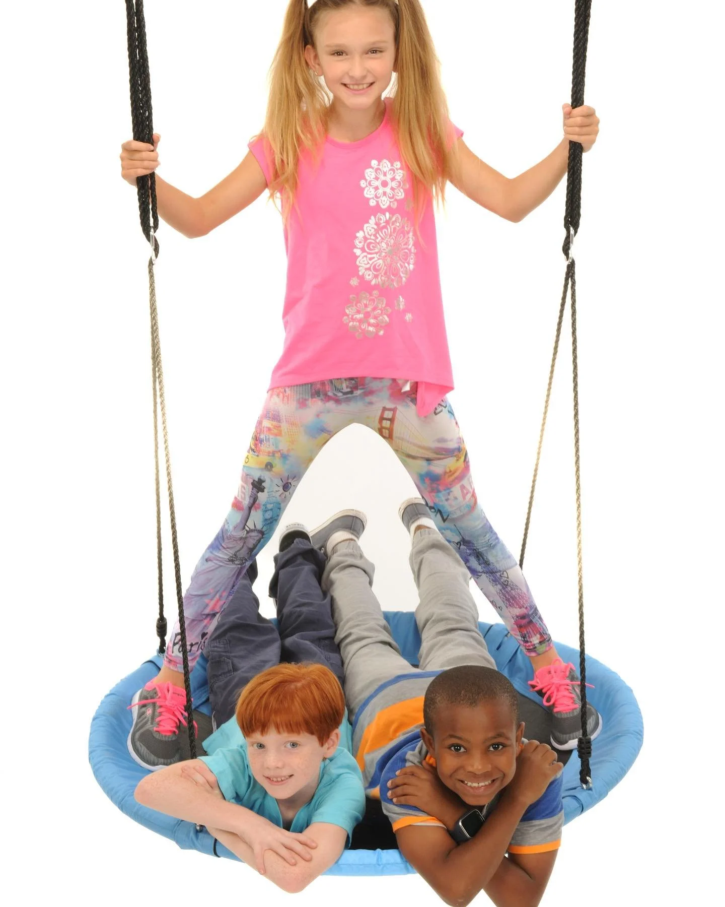 Four children playing on a circular swing, with a girl standing on the swing and the three boys lying inside it, smiling.