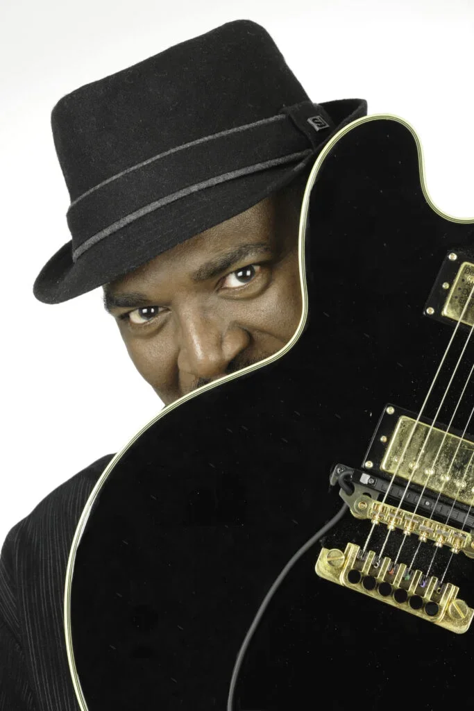 A man wearing a black hat peeking from behind a black guitar with gold hardware.