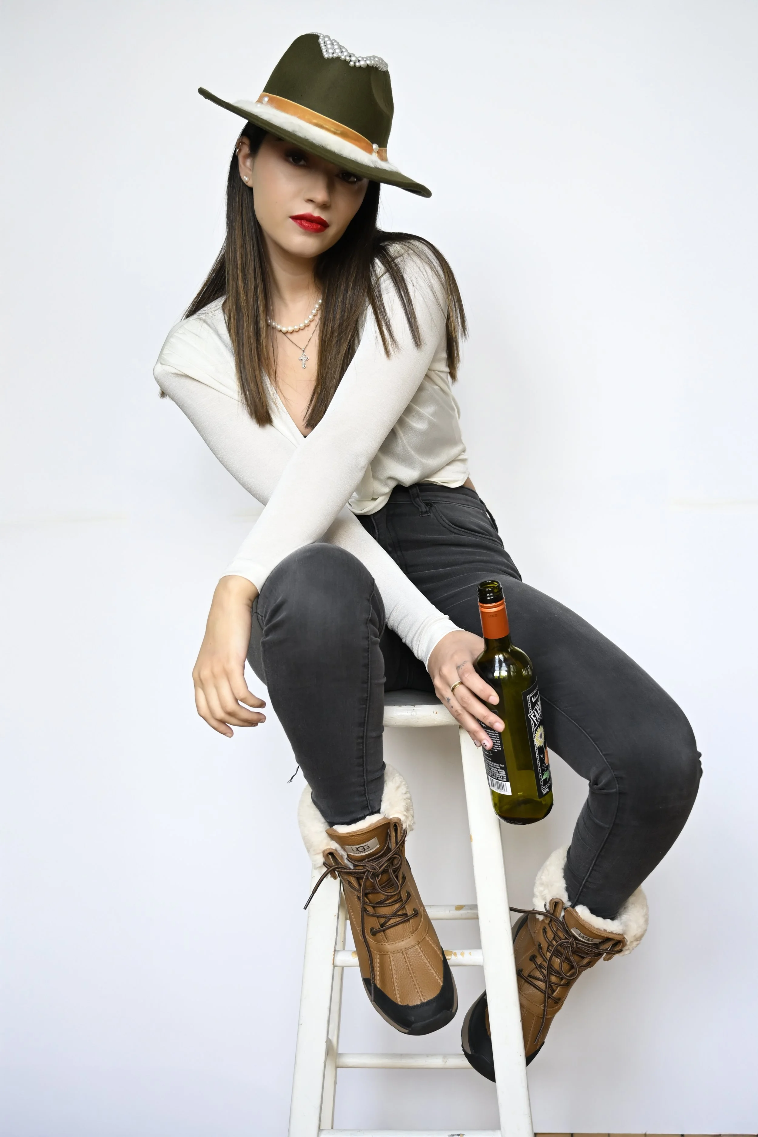Woman sitting on a white ladder holding a beer bottle, wearing tan winter boots, black jeans, a white long-sleeve shirt, red lipstick, jewelry, and a green wide-brimmed hat with decorative pearls and a ribbon.