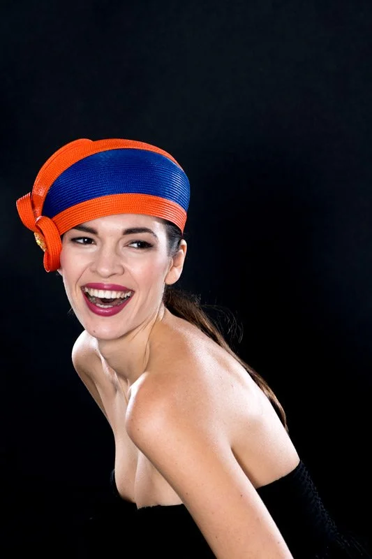 A woman with dark hair wearing a colorful orange and blue headwrap, smiling against a black background.