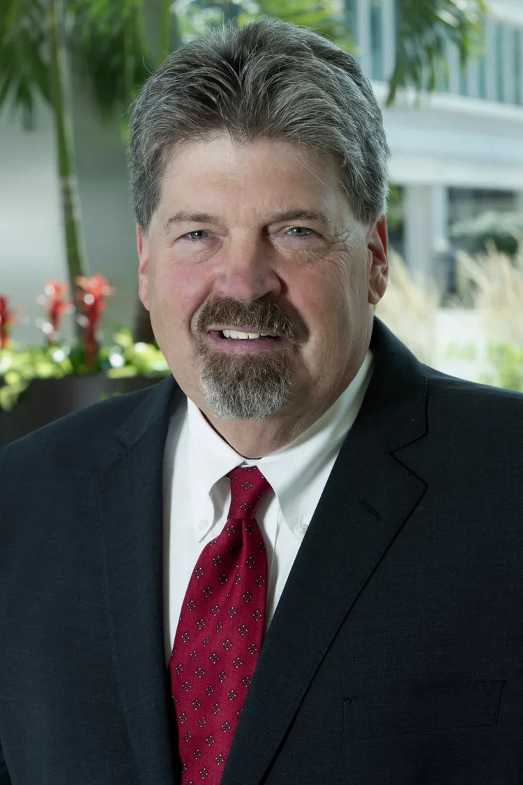 A middle-aged man with light skin, gray hair, and a beard wearing a dark suit, white shirt, and red patterned tie, standing outdoors with blurred greenery and modern building in the background.