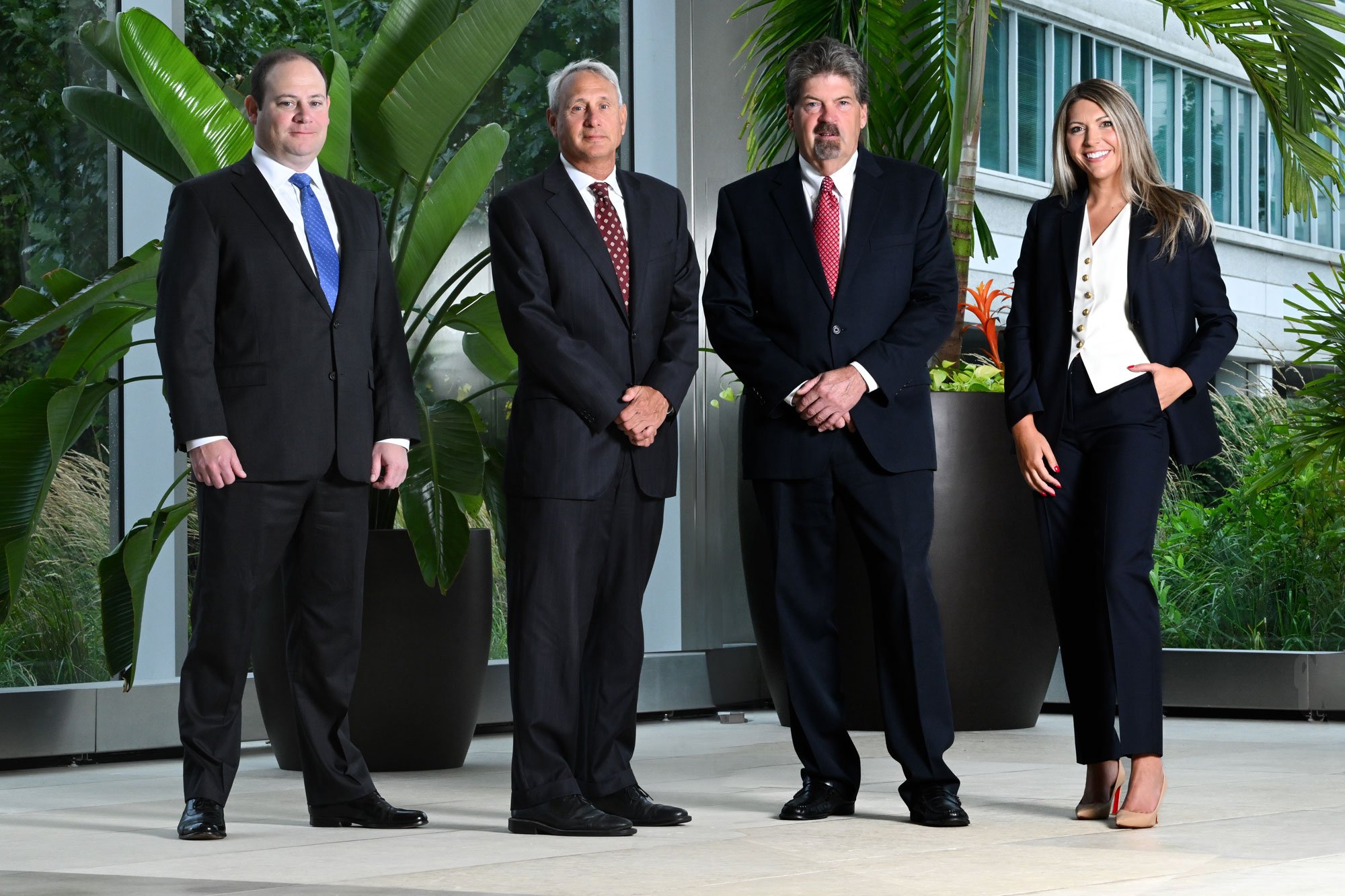 Four professionally dressed people, three men and one woman, standing side by side in an indoor space with large green plants and a glass window in the background.