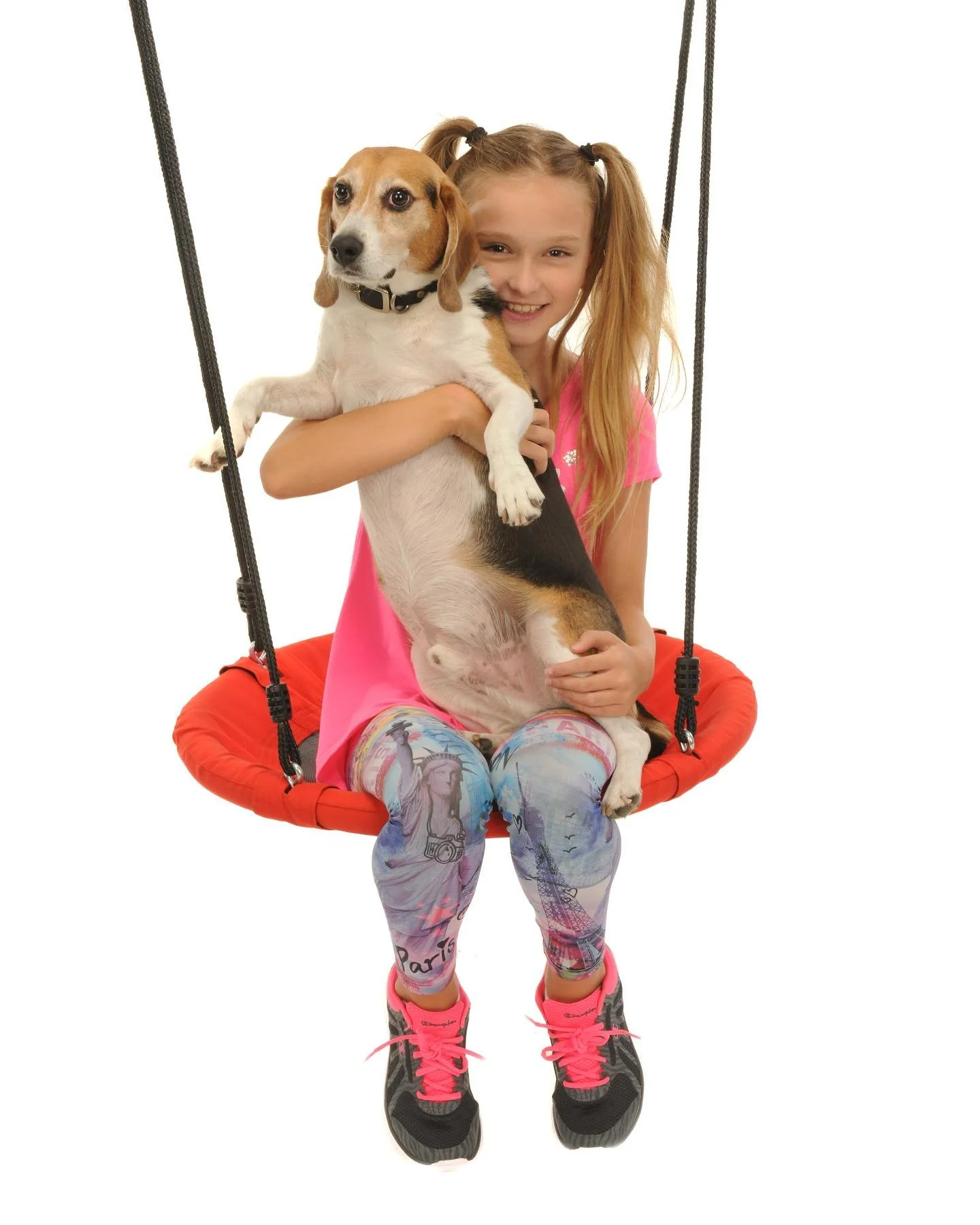 A young girl with blonde hair in pigtails sitting on a red swing, holding a beagle dog with a black, white, and brown coat.