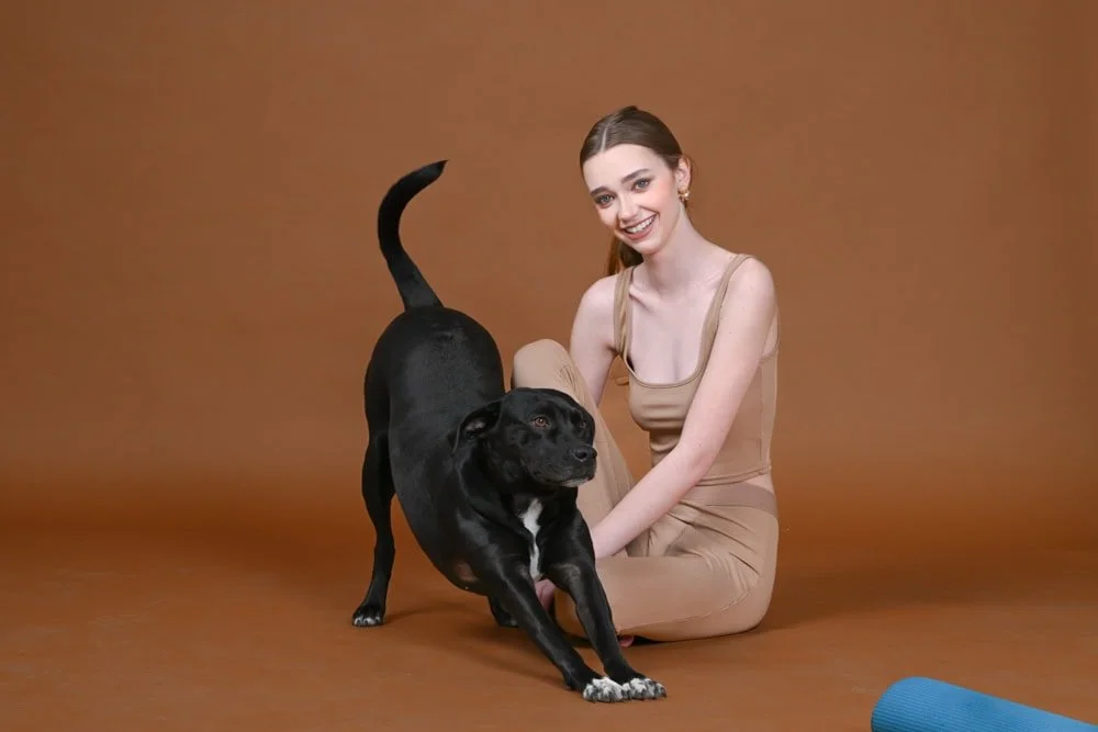A young woman with long brown hair, wearing a beige sleeveless top and matching pants, sitting on her knees on a brown background, smiling while playing with a black dog, possibly stretching or exercising.