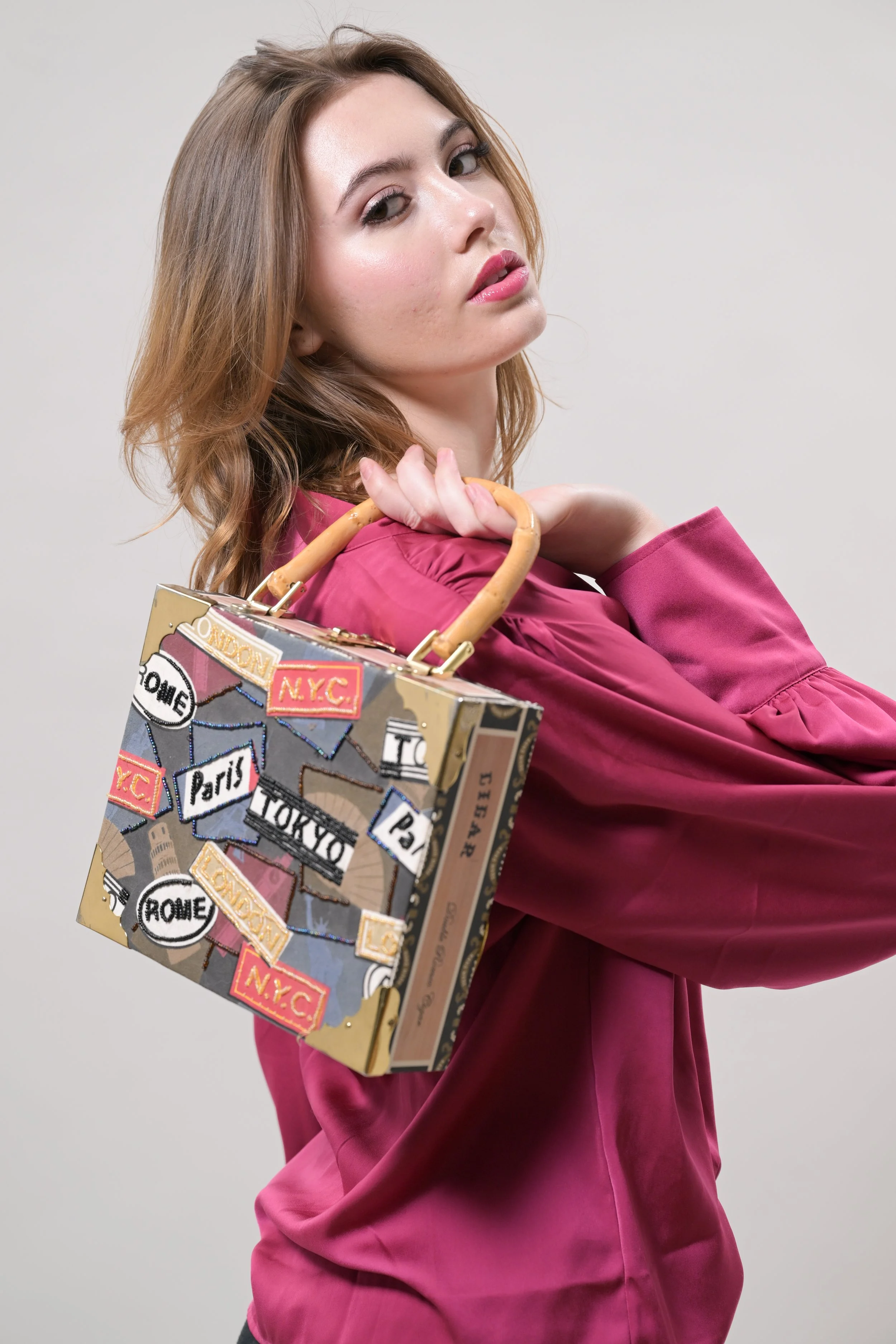 A woman with light skin and wavy brown hair wearing a pink satin blouse, holding a colorful patterned bag with city labels like NYC, Paris, Tokyo, and Rome over her shoulder.