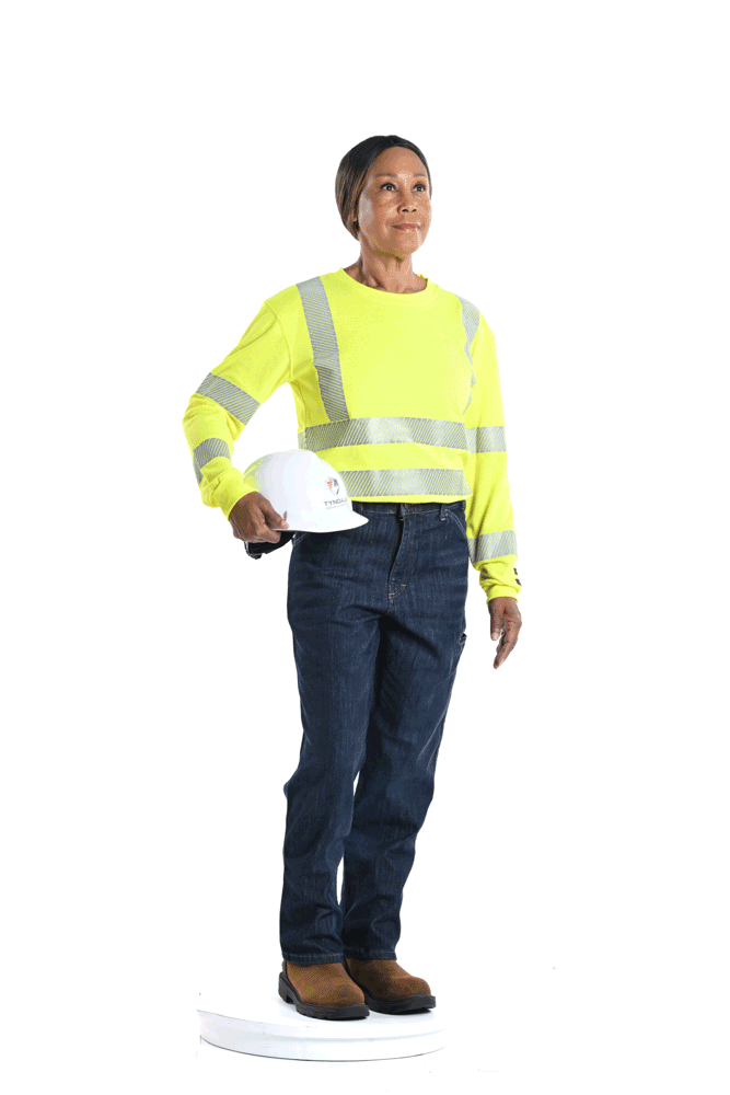 A woman in a yellow safety high-visibility shirt with reflective stripes, dark blue jeans, brown boots, holding a white safety helmet in her right hand, standing on a circular white platform.