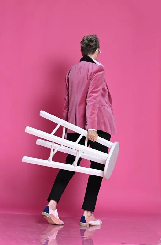 Person in pink blazer walking away against a pink background, carrying a white wooden chair.