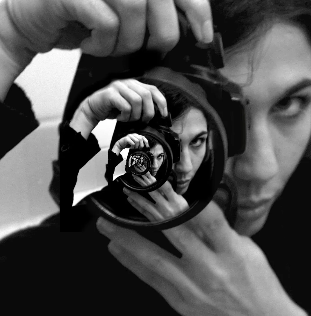 A woman taking a black and white photo of herself with a camera, creating multiple nested reflections through the camera lens.