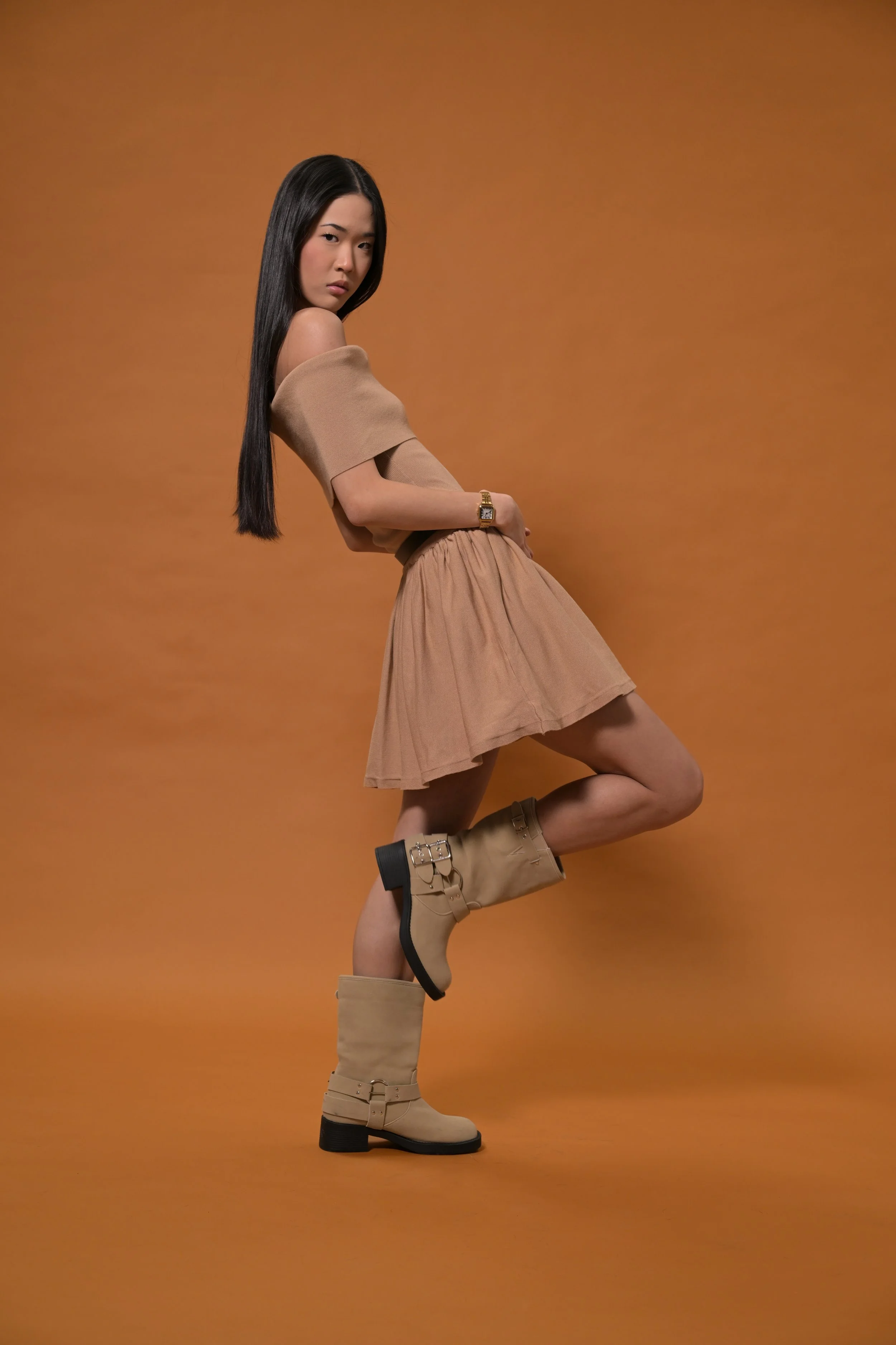A woman with long dark hair wearing a tan off-shoulder top, a tan skirt, and tan boots, standing against an orange background.