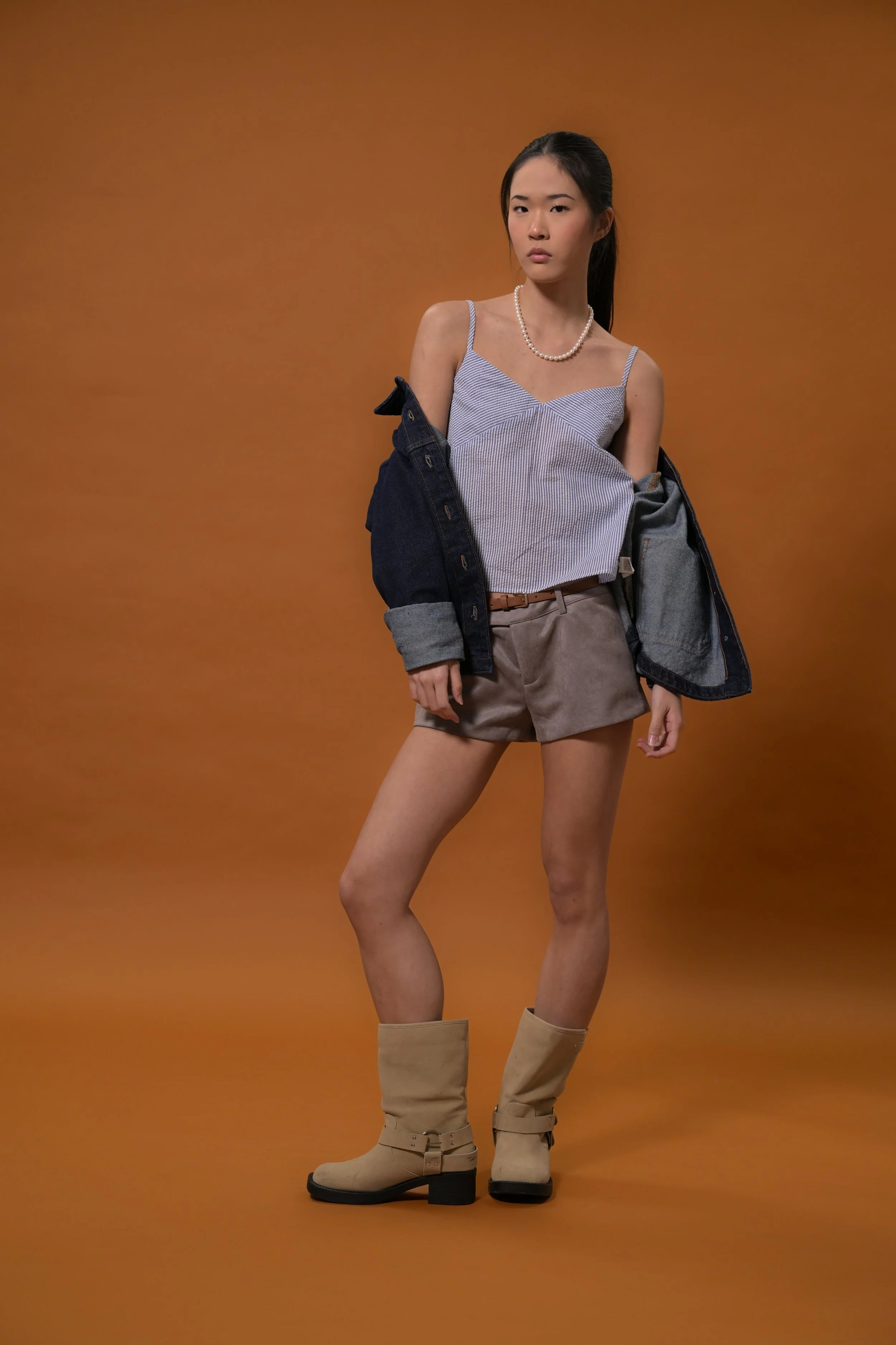 Young woman in beige boots, gray shorts, blue striped tank top, black jacket, and pearl necklace posing against a brown background.