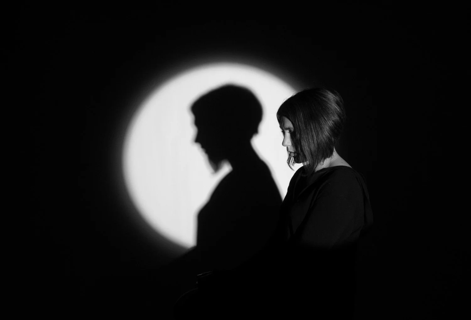 Black and white photo of a woman sitting with her head bowed, facing right, with her portrait's shadow cast behind her on a white circular background.