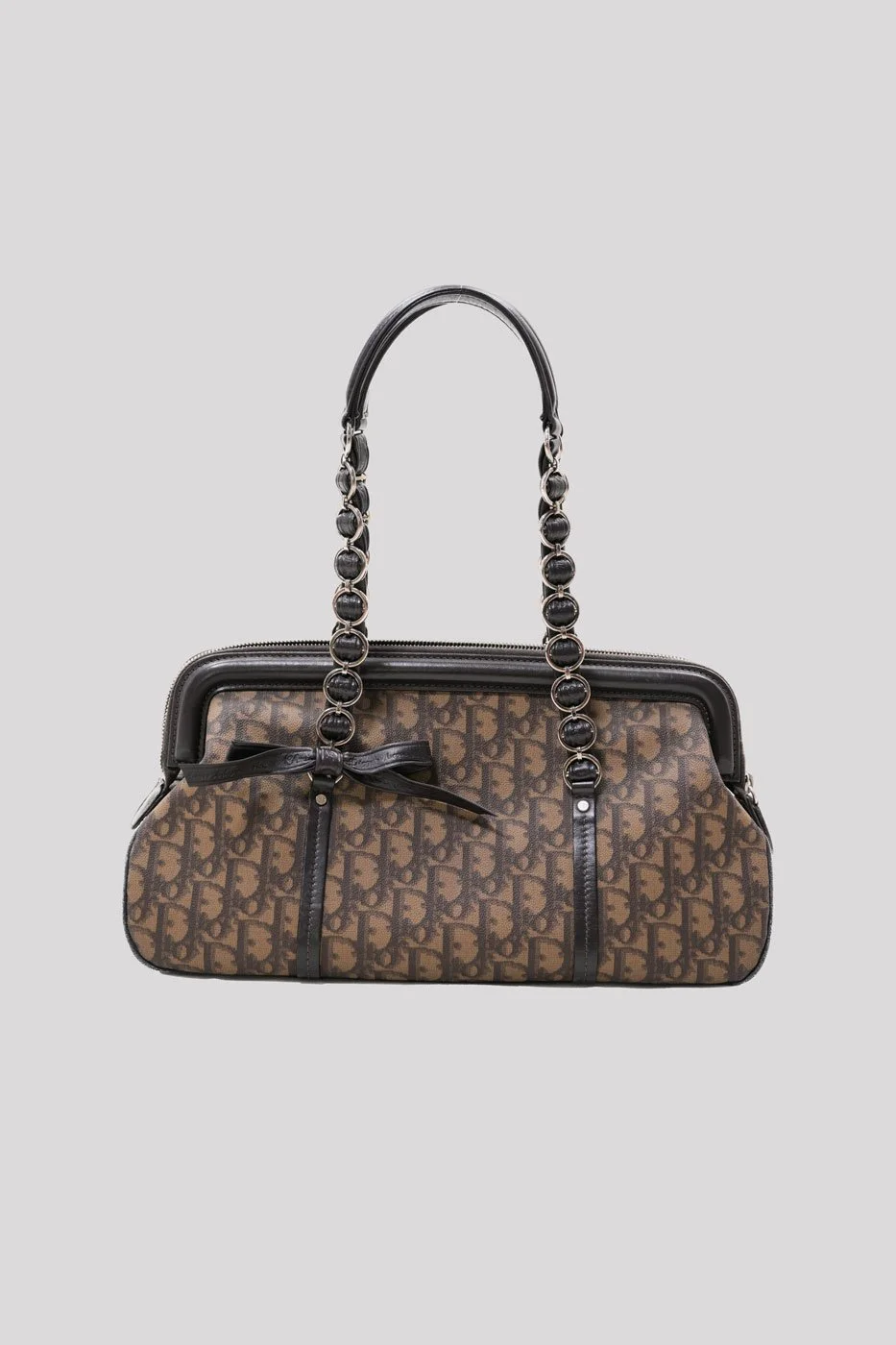 A designer handbag with a brown monogram pattern, black leather handles with decorative metal rings, and black leather accents, featuring a decorative bow on the side.