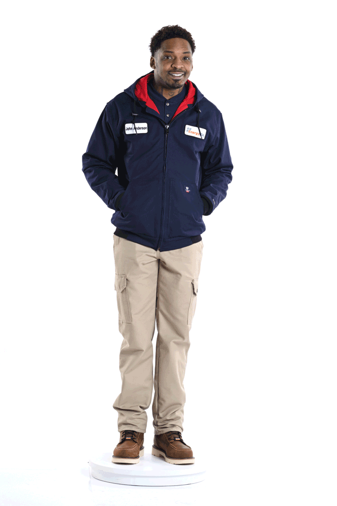 A man with short curly hair and a beard standing on a white platform, wearing a navy blue jacket, beige cargo pants, and brown boots, smiling at the camera.