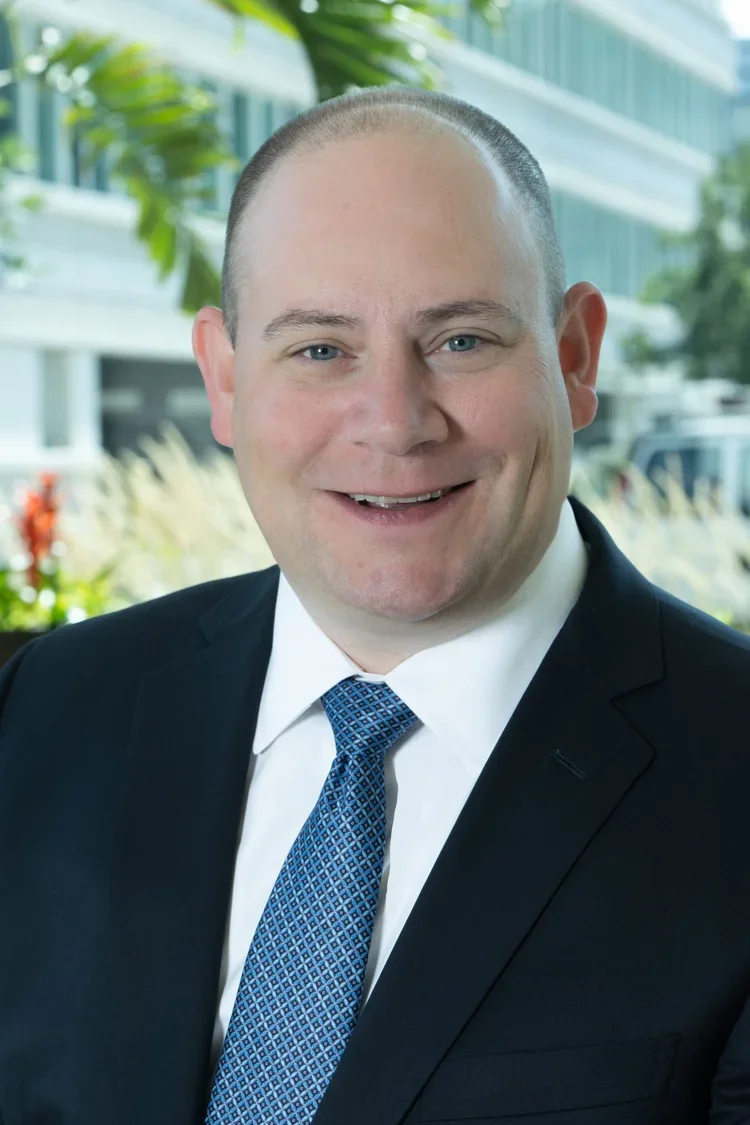 Headshot of a smiling man in a suit and tie outdoors with a blurred building and greenery in the background.