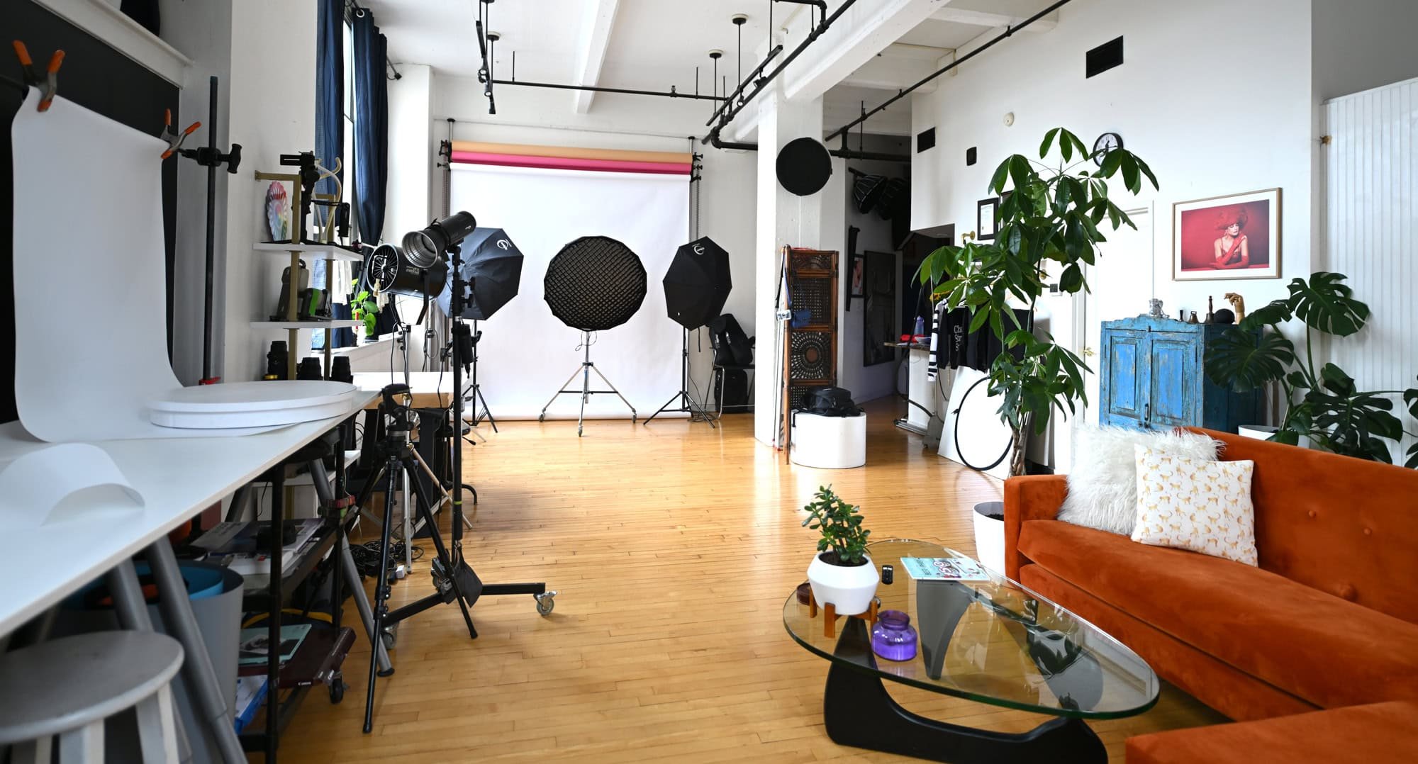 A photography studio with a white backdrop and studio lighting equipment, including umbrellas and softboxes. There are wooden floors, a blue cabinet, and a cozy seating area with an orange couch, pillows, and a glass coffee table. The room features large plants, framed artwork on the wall, and various photography tools and accessories.