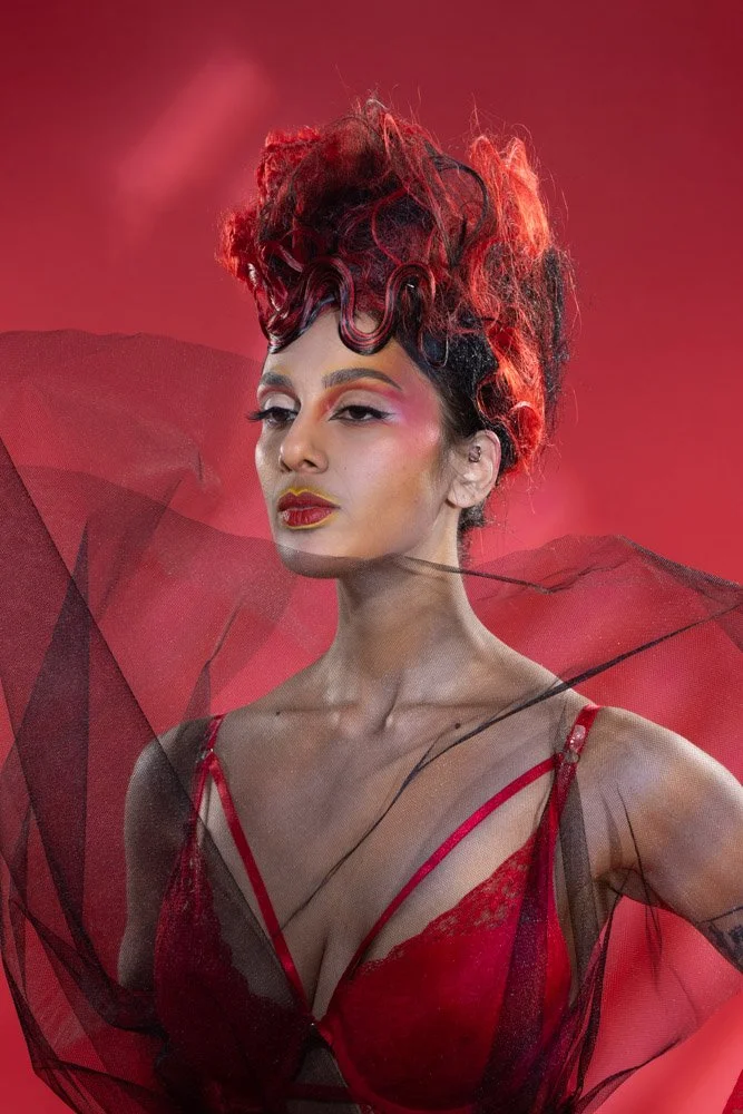 A woman with elaborate red and black curly hair, wearing dramatic makeup, red lipstick, a red lace bra, and black sheer fabric, posing against a red background with sheer fabric accents.