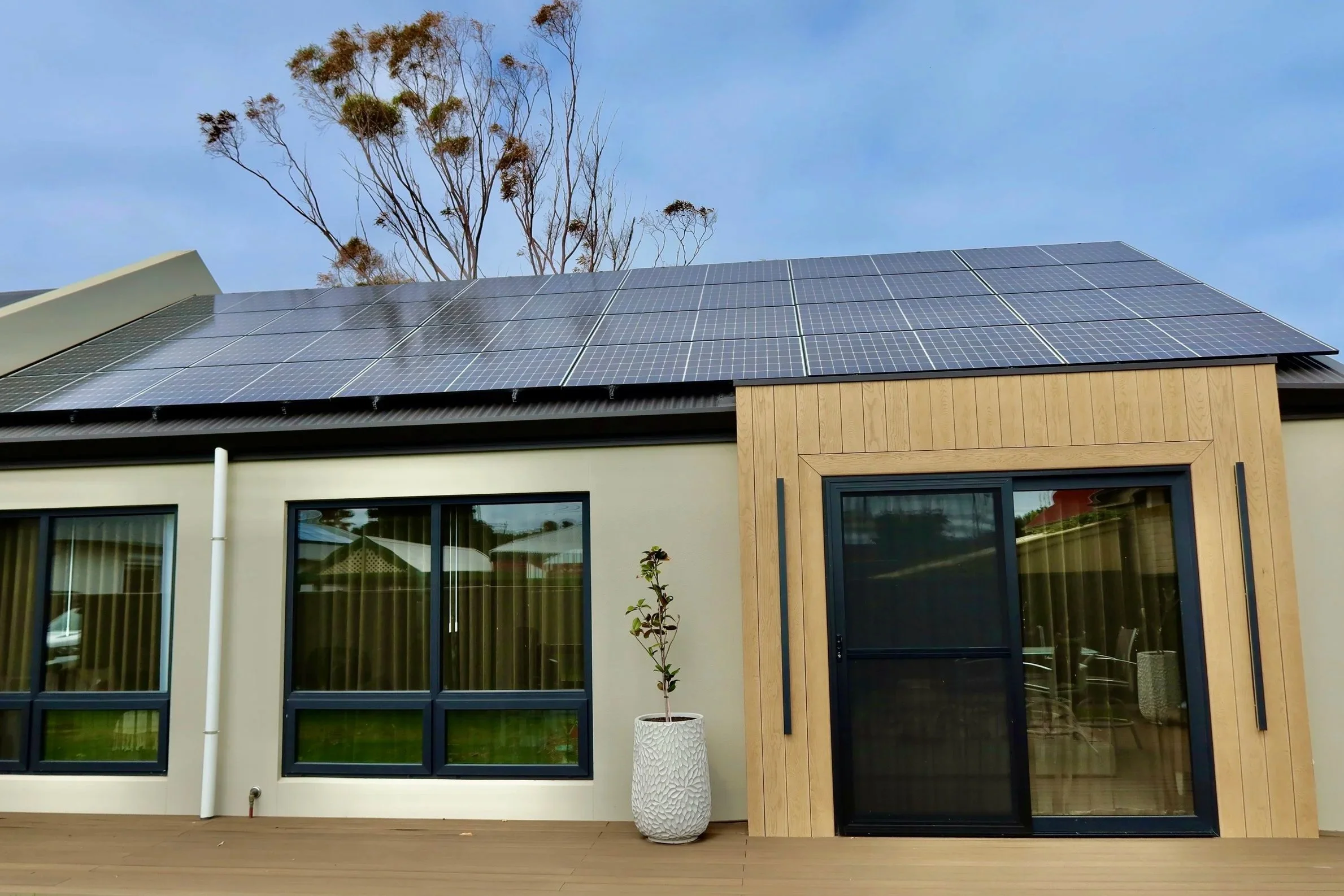 Modern house with solar panels on the roof, large windows, a sliding glass door, a wooden accent wall, potted plant, and a wooden deck, with trees and a blue sky in the background.