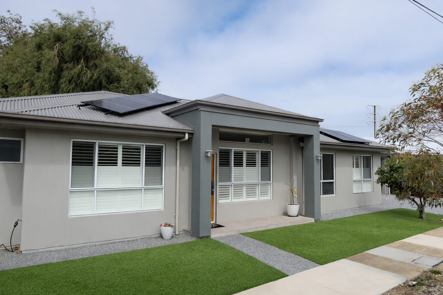 Modern single-story house with gray exterior, large windows with white shutters, a covered front porch, and solar panels on the metal roof. The front yard has a green lawn with small plants, flowers, and trees.