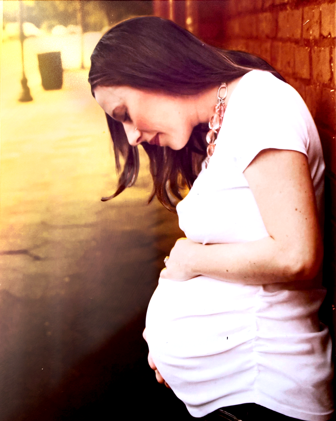 A pregnant woman with long brown hair wearing a white shirt and necklace, leaning against a brick wall, looking down with a thoughtful expression.