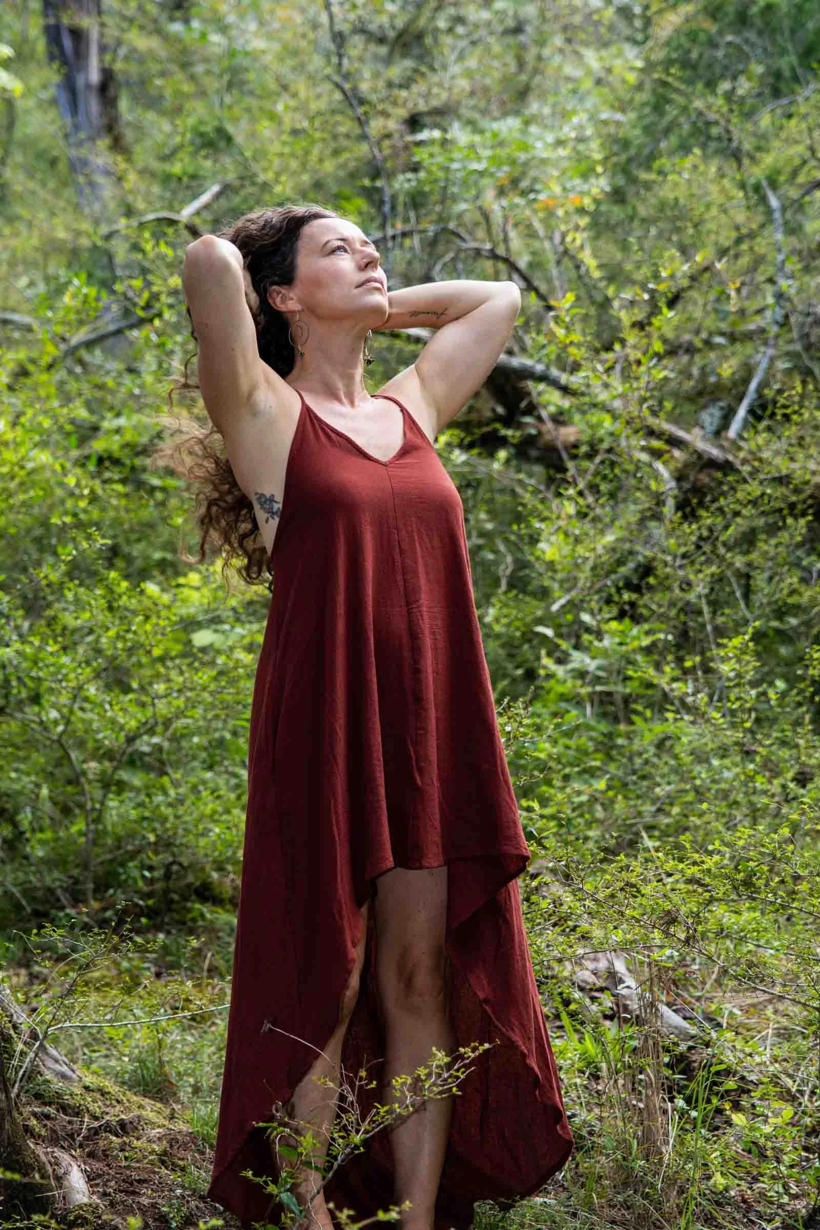A woman in a red sleeveless dress standing in a green forest with arms raised behind her head, looking upward.