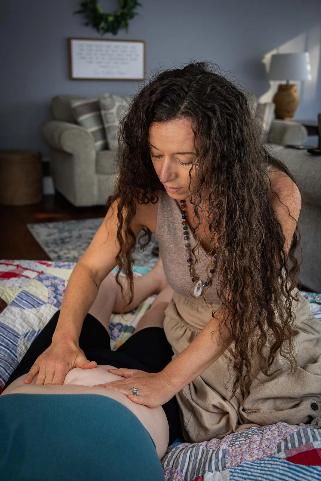 A woman practicing massage therapy on a person's leg in a cozy living room with furniture and wall decorations.