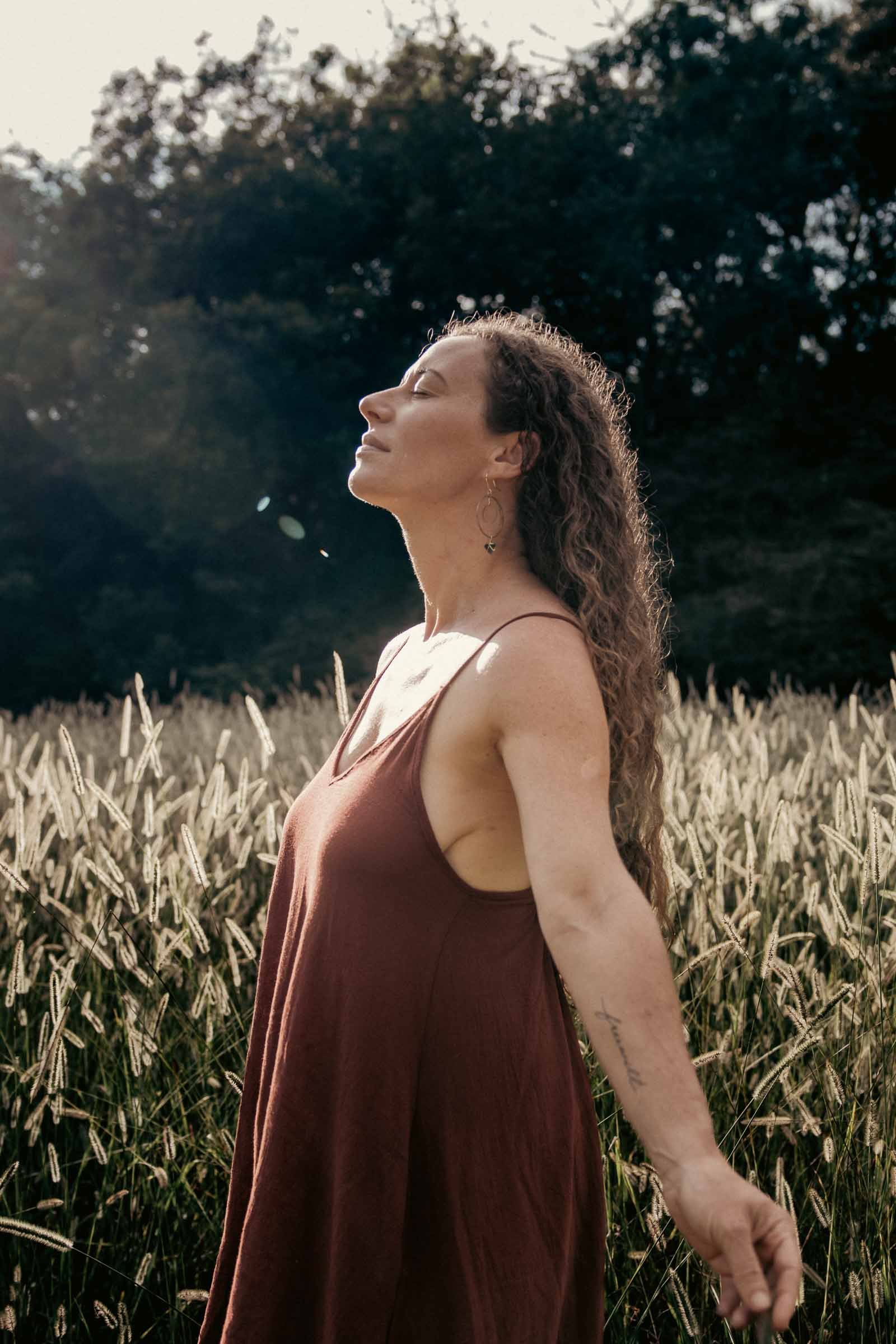 A woman with long, curly hair standing in a wheat field with her eyes closed and face tilted upward as she enjoys the sunlight.