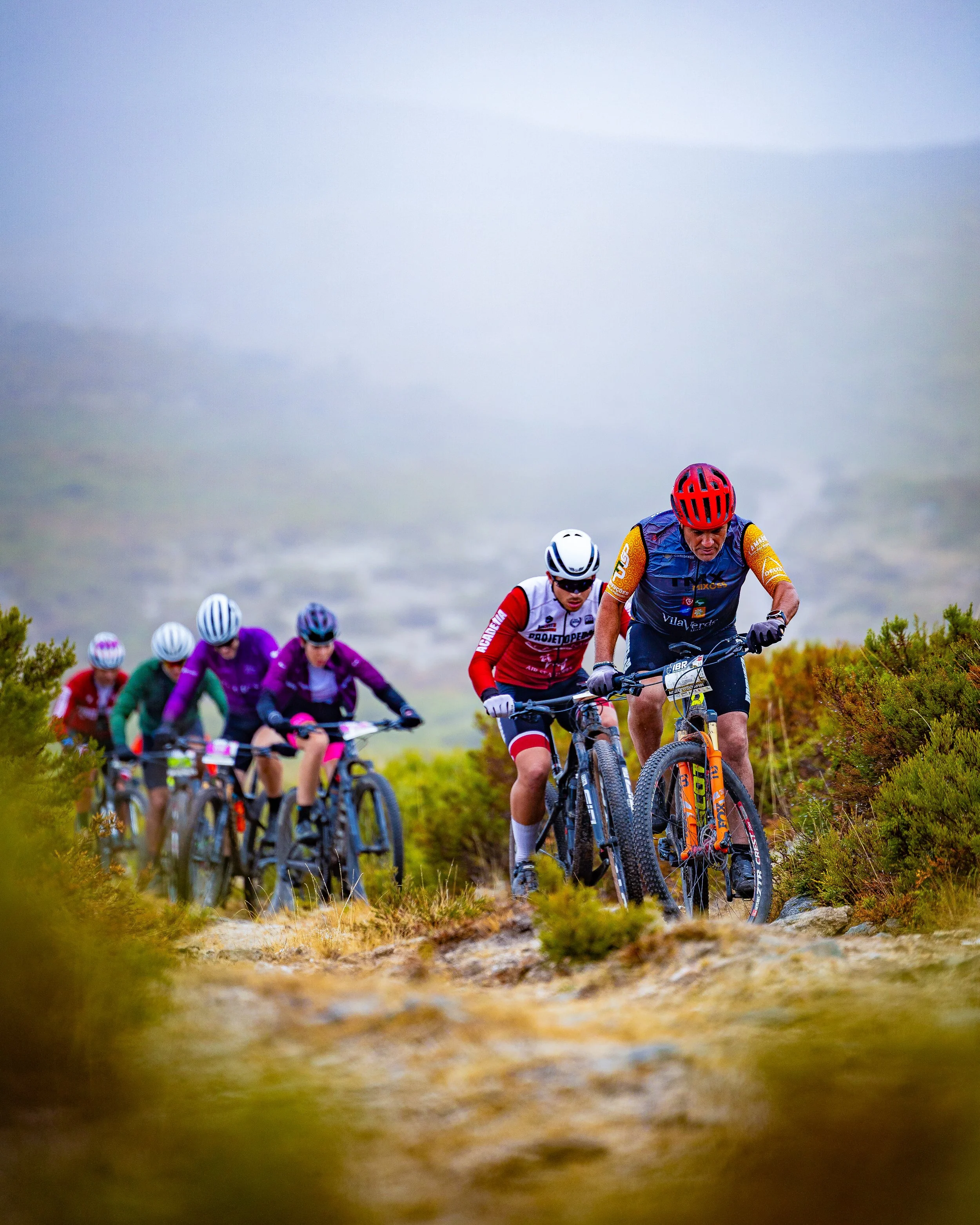 A group of mountain bikers wearing helmets and sports gear riding on a trail through a hilly landscape with greenery.