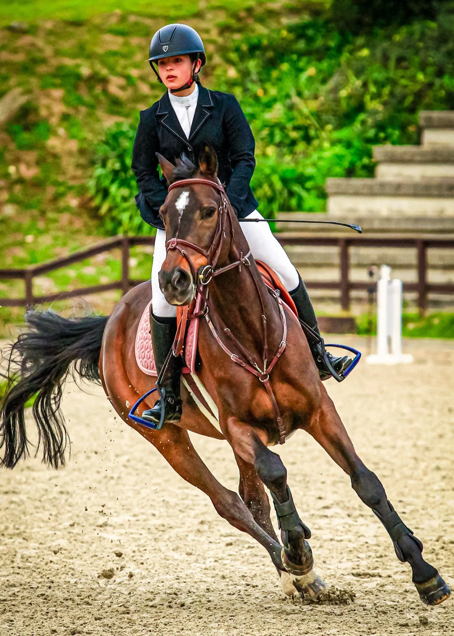 A young female equestrian wearing a helmet and riding attire, riding a brown horse with a pink saddle pad in an outdoor riding arena with green foliage and stairs in the background.