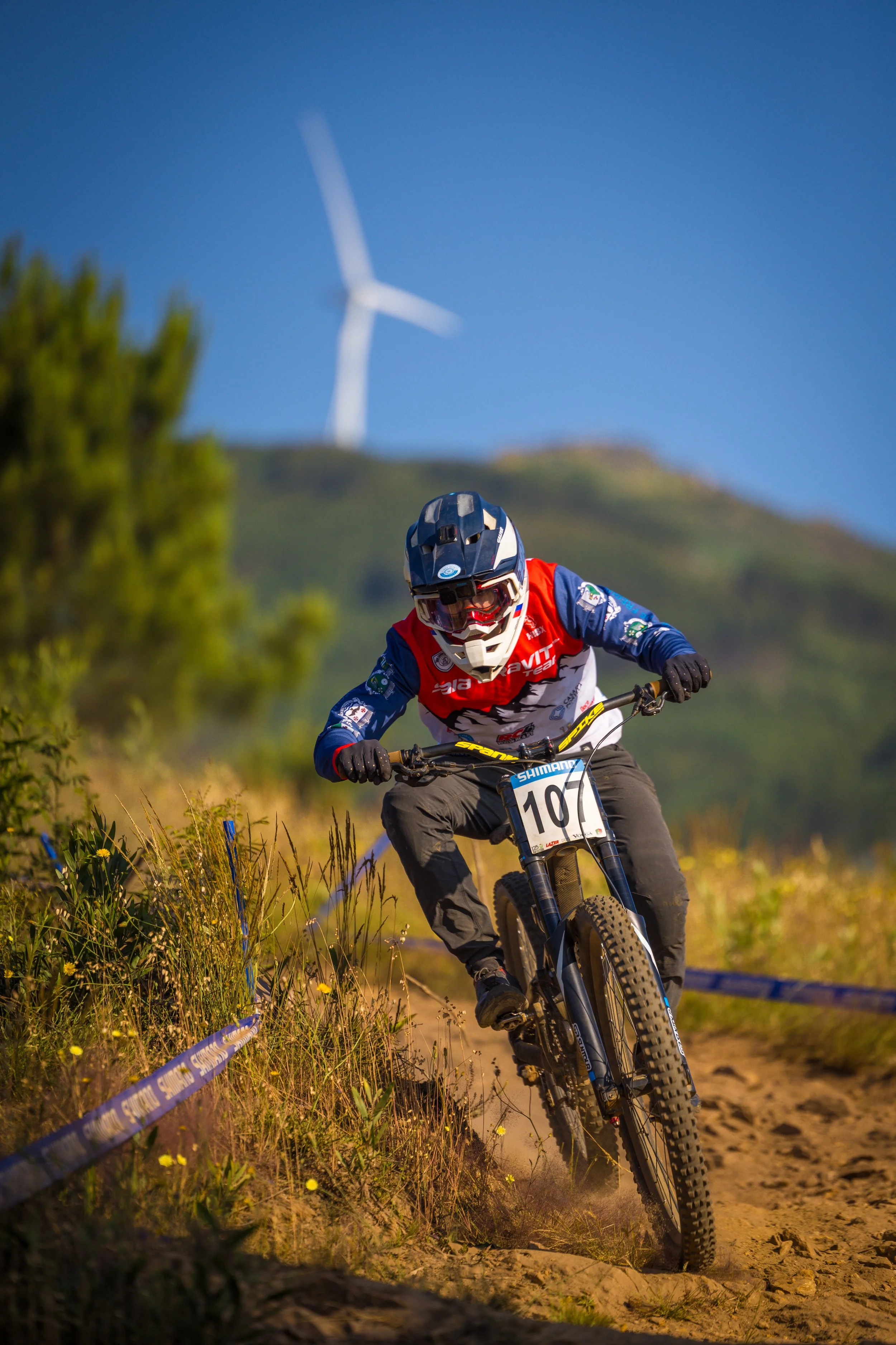 A mountain biker wearing a helmet, goggles, and a red and blue jersey riding downhill on a dirt trail with plants and a wind turbine in the background.