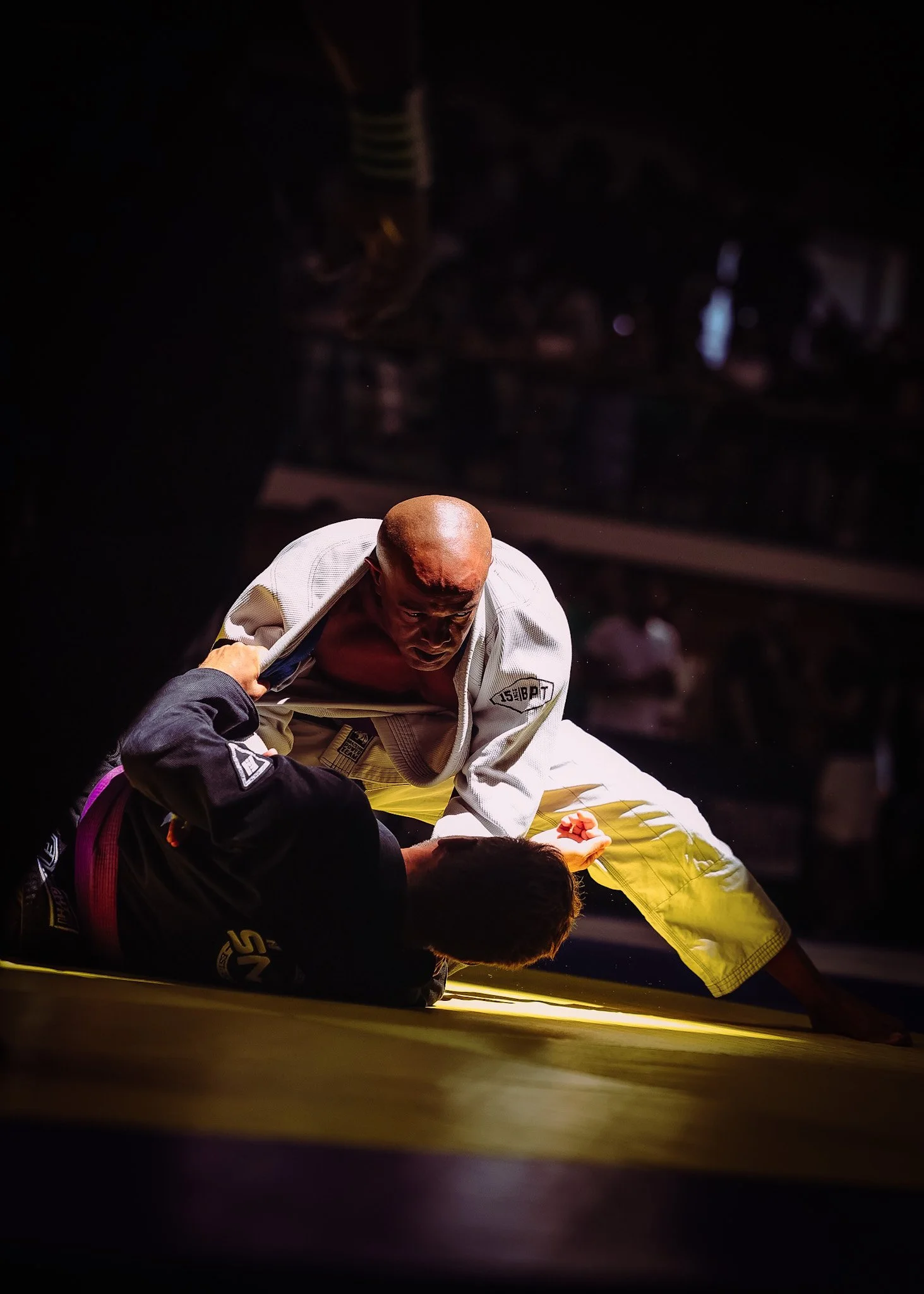 Two men practicing Brazilian Jiu-Jitsu on a yellow mat, one in a white gi and the other in a black gi, with a dark background.