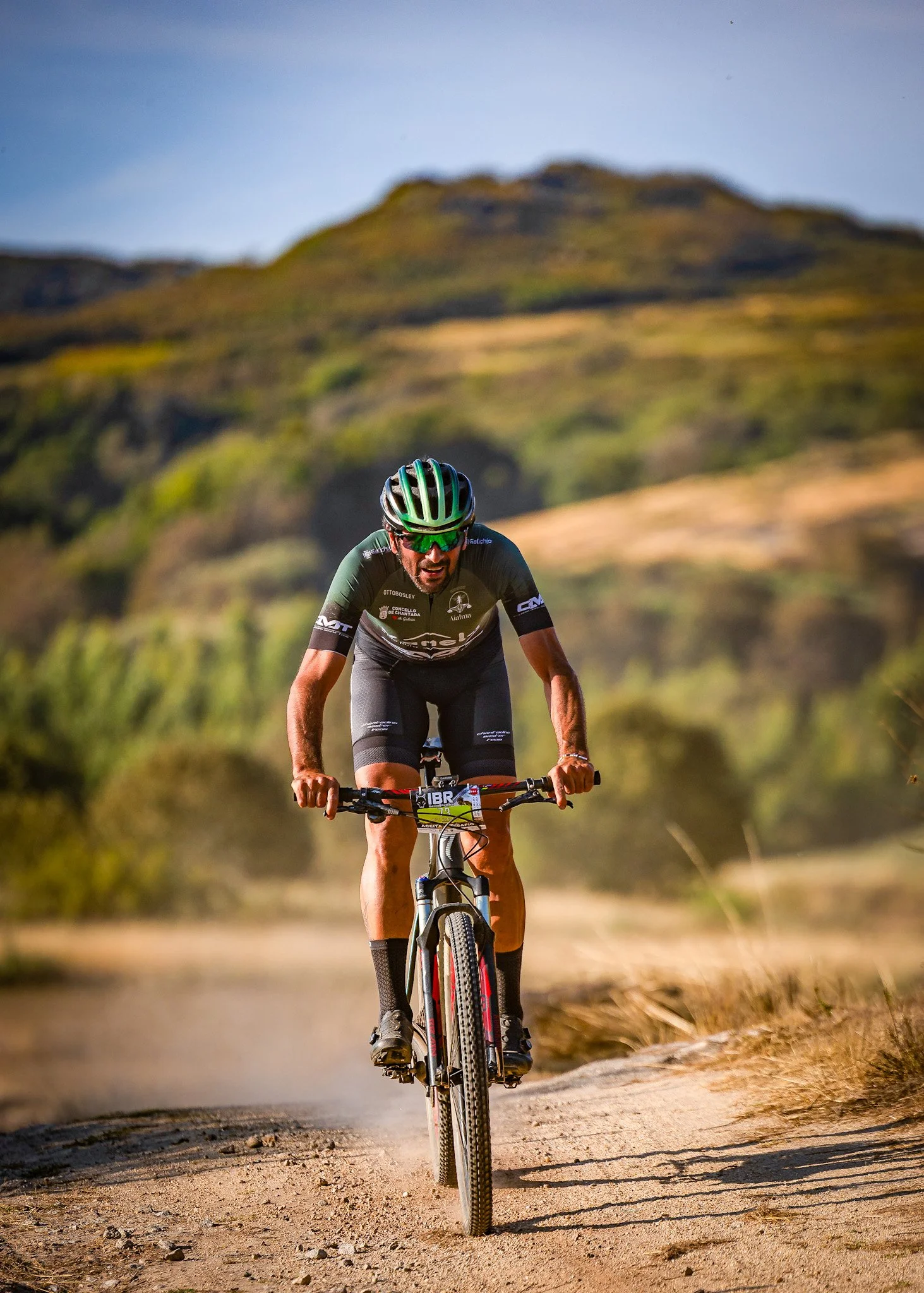 A man riding a mountain bike on a dirt trail in a hilly outdoor landscape under a clear sky.