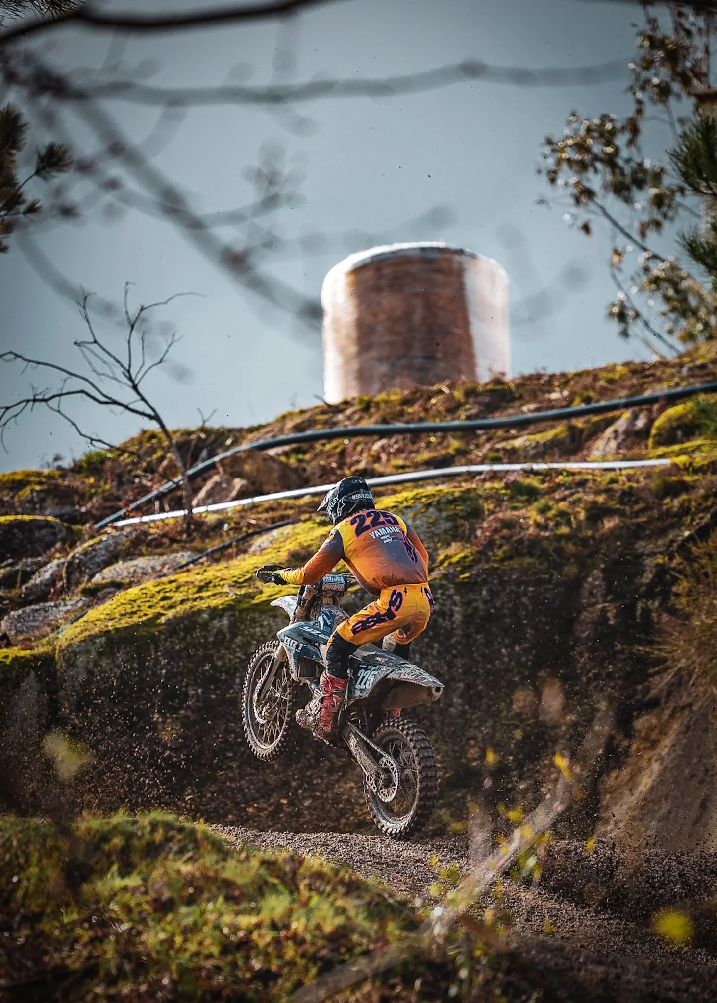 A motocross rider in orange and purple gear riding a dirt bike through a muddy trail outdoors, with trees and a cloudy sky in the background.
