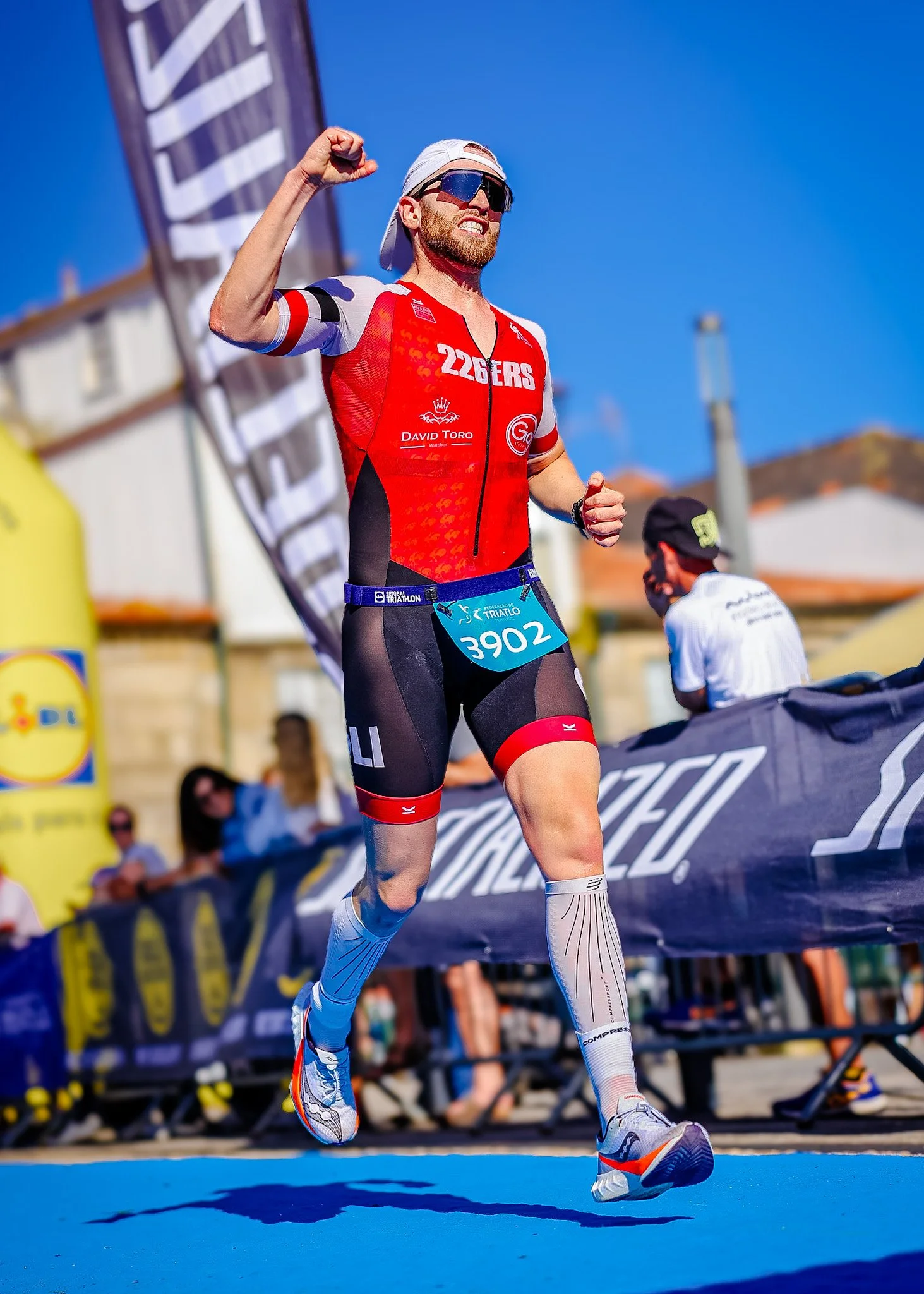Male triathlete crossing the finish line during a race, wearing a red and black uniform, sunglasses, and a white cap, with arms raised in celebration.