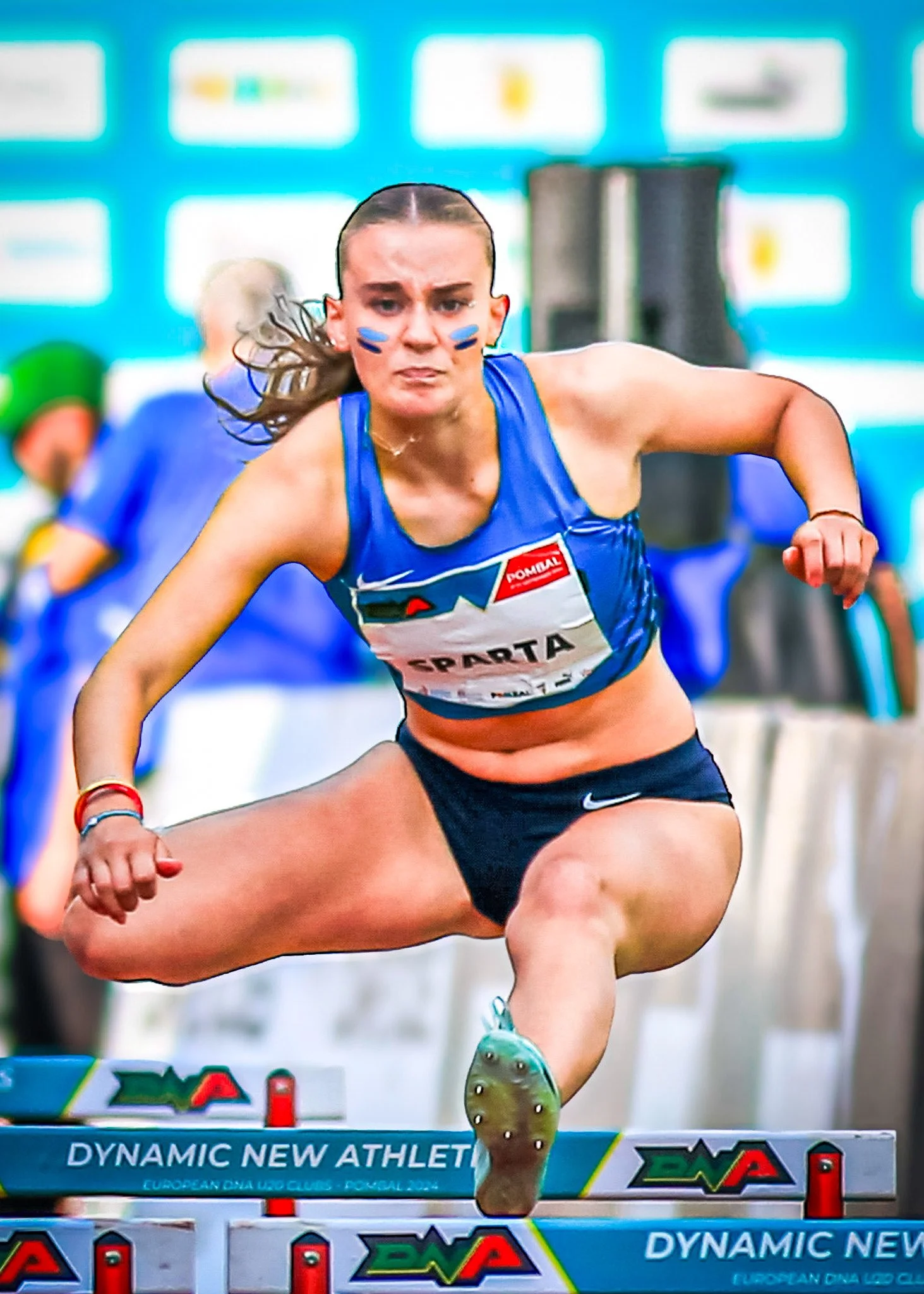 Female athlete in blue sports bra and black shorts jumping over a hurdle during a track and field race.