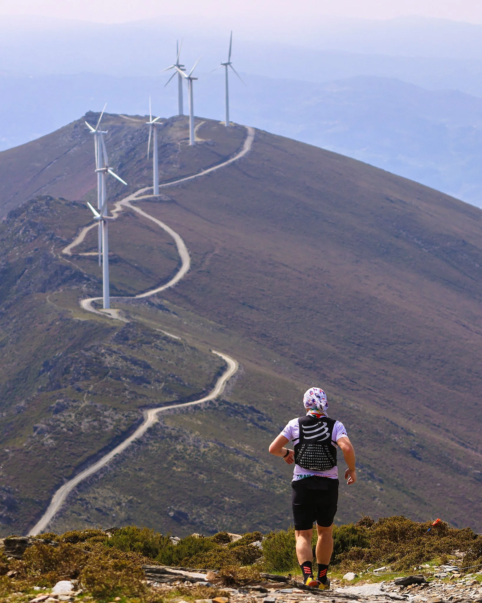 A person running uphill on a trail near bushes with wind turbines on a mountain in the background.