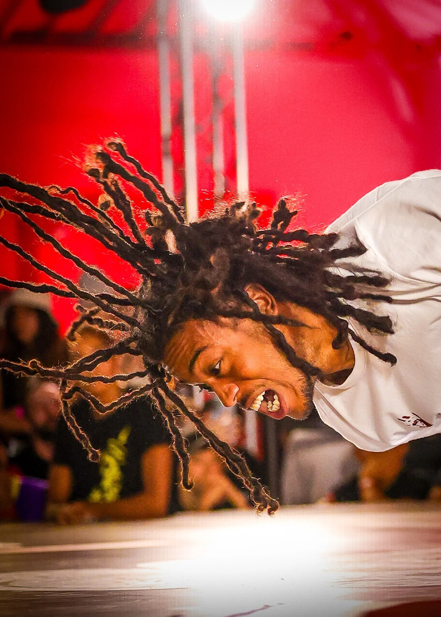 A man with dreadlocks in the air, leaning forward with an intense expression, in a martial arts or dance studio with a red wall in the background.
