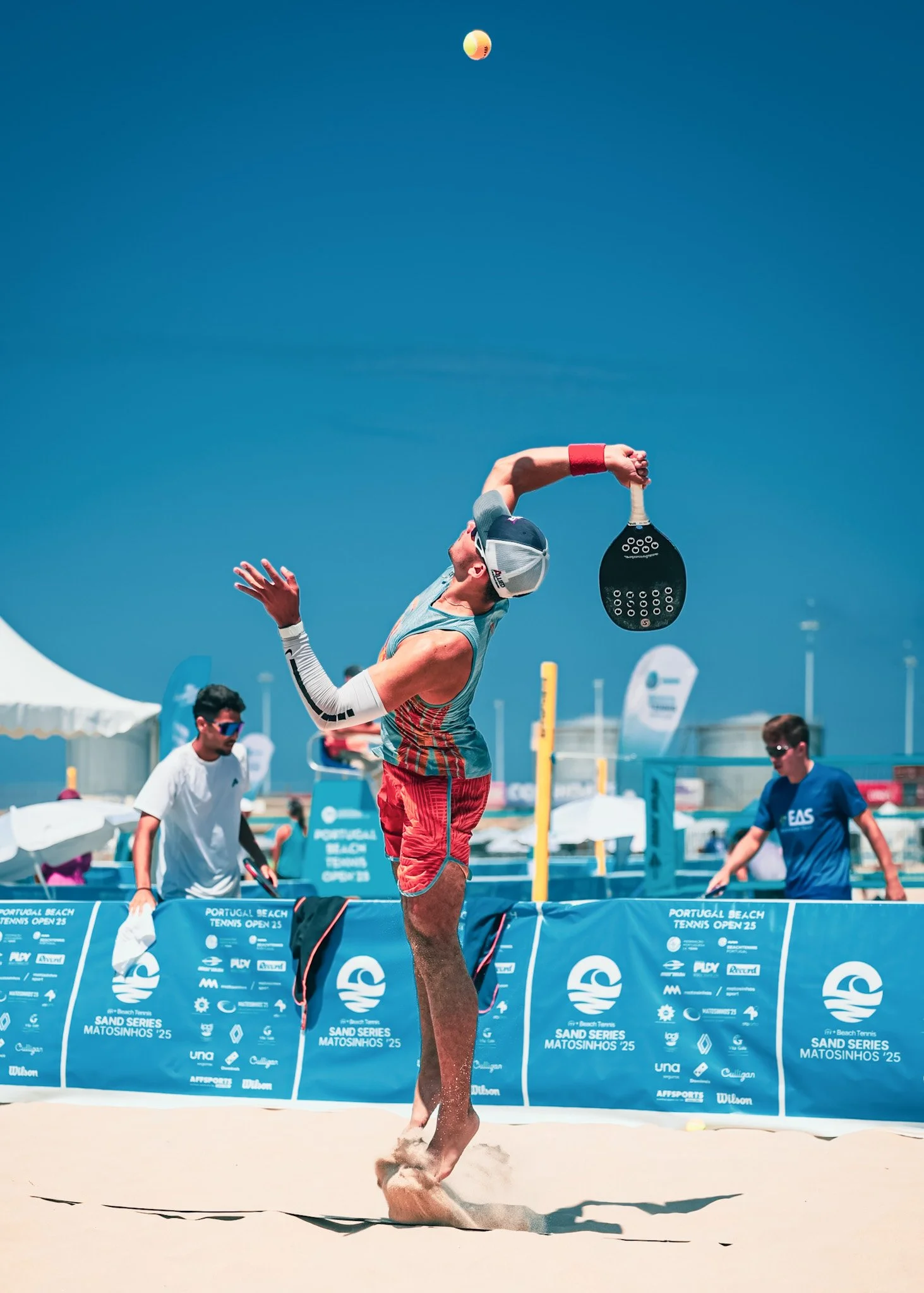A man playing beach tennis on a sandy court during daytime under a clear blue sky, with other players and banners in the background.