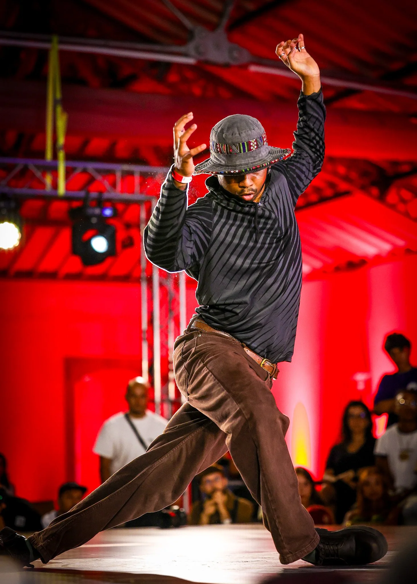 Person dancing on stage in a black and gray striped jacket and brown pants, wearing a bucket hat, with a red lit background and audience watching.