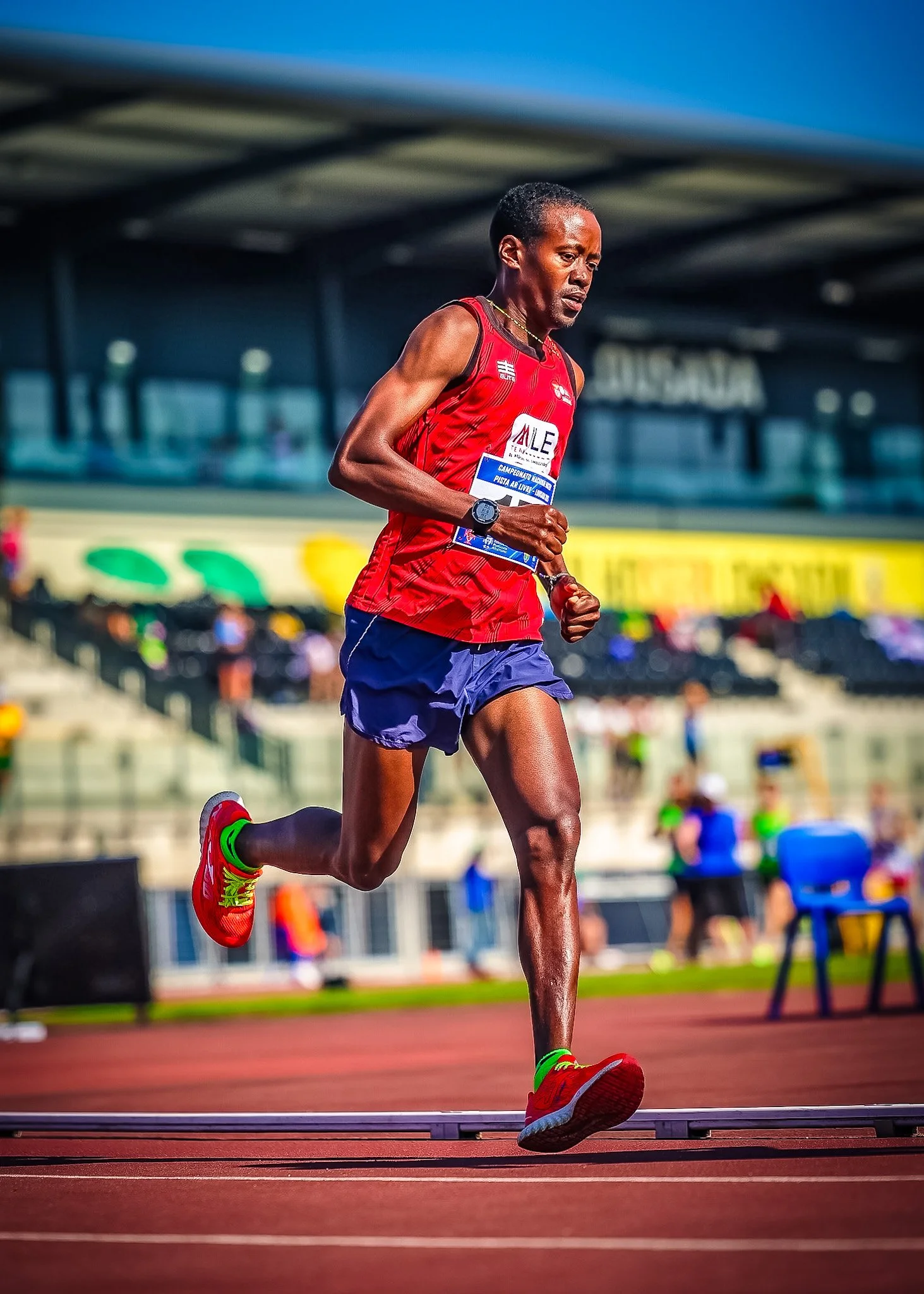 A male marathon runner in red tank top, blue shorts, and red running shoes, running on a track at a stadium during a race.