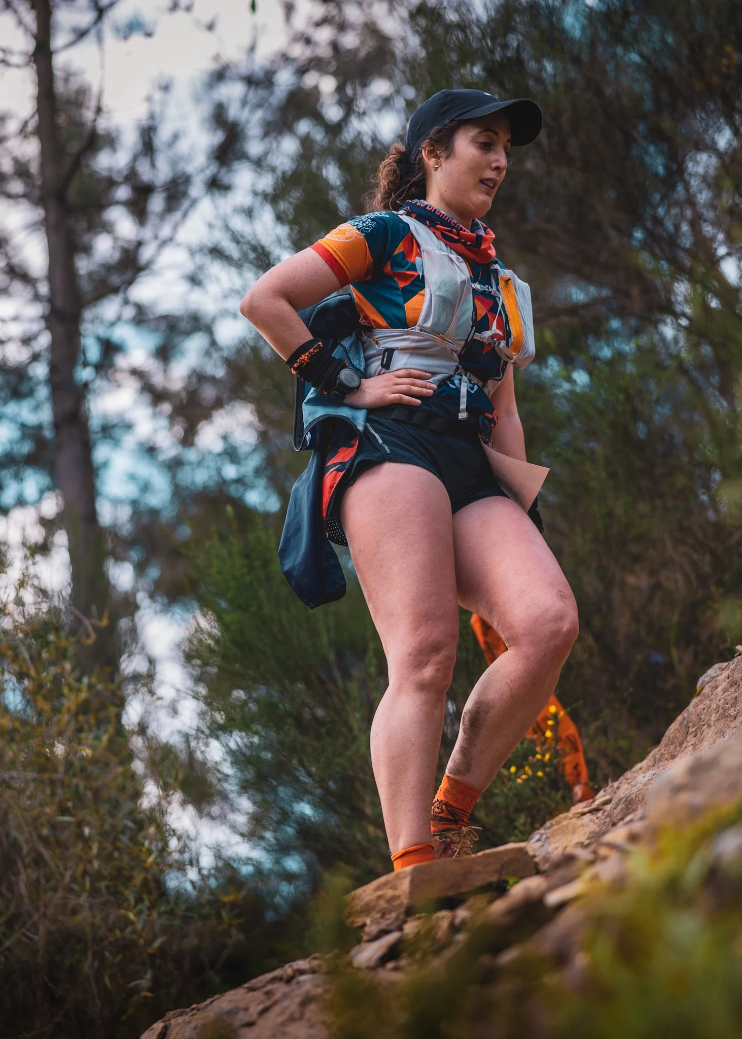 Female trail runner in athletic gear, climbing rocky terrain outdoors with trees and sky in background.