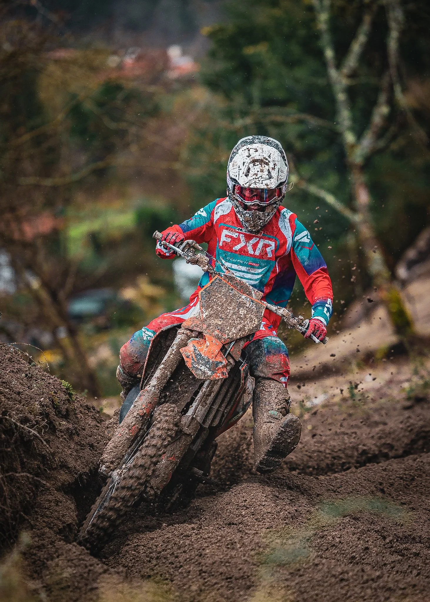 A motocross rider in a red, blue, and white racing suit and helmet riding a muddy dirt trail on a mountain bike or motorcycle in a forested area.
