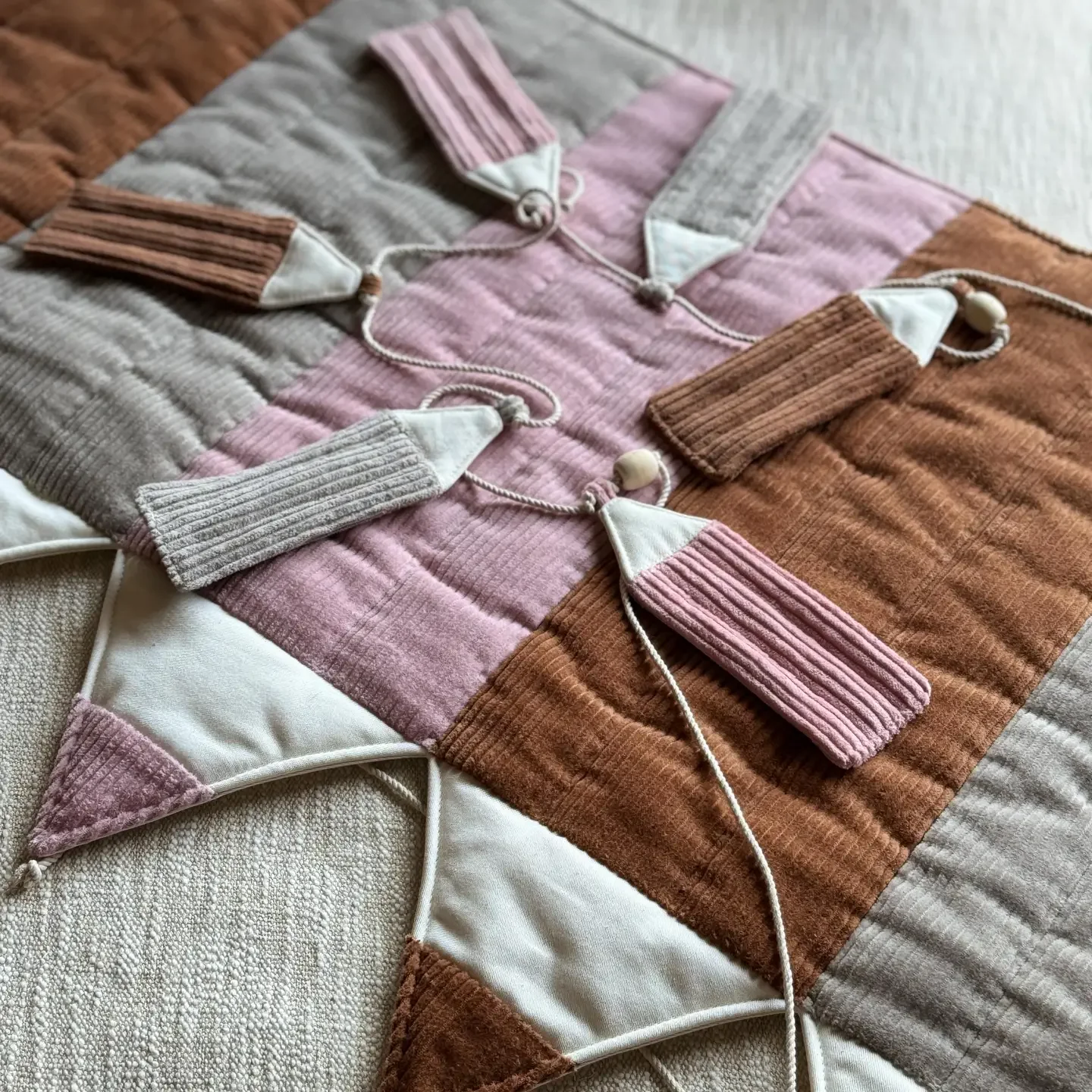 Close-up of a quilted textile wall hanging with knitted sock-like decorations hanging from cords in shades of pink, brown, gray, and cream.