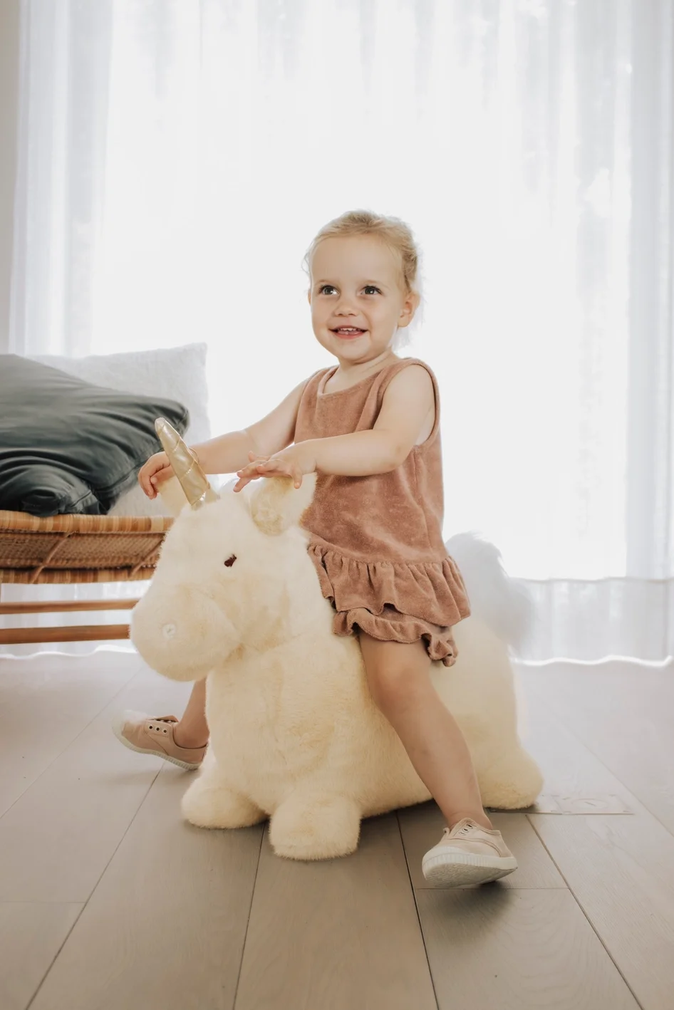 A young girl with blonde hair riding a plush unicorn toy in a bright room, wearing a light brown sleeveless dress and white shoes.